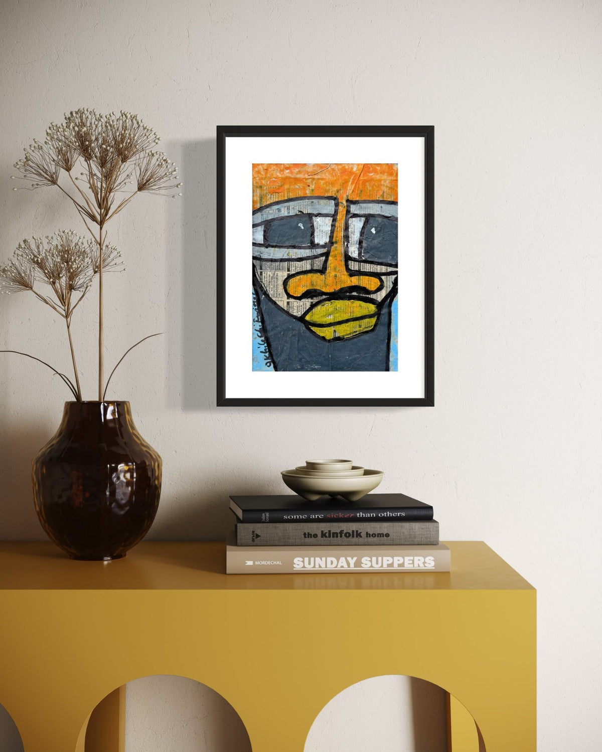 A framed art print featuring an abstract face illustration in orange, yellow, blue, and gray, displayed on a white wall above a yellow console table with books and a vase.