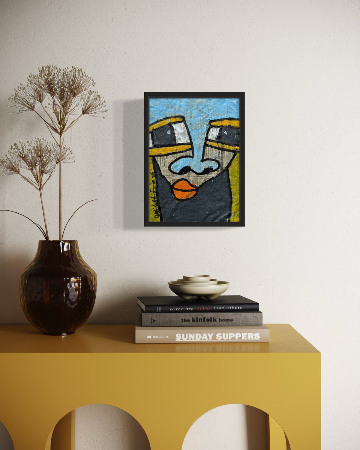 A framed abstract art print featuring a stylized face in blue, yellow, black, and orange, displayed on a white wall above a yellow console table with a dark brown vase and books.