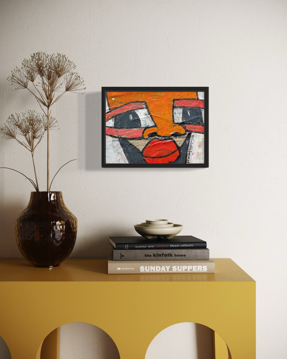 A framed abstract art print featuring a stylized face in orange, red, black, and white, displayed on a white wall above a yellow console table with a dark brown vase and books.