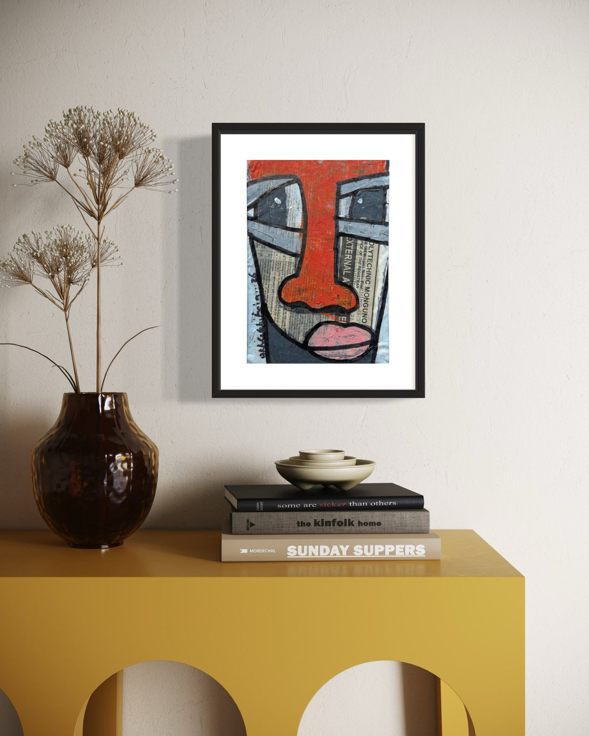 A framed art print featuring an abstract face with a prominent red nose hangs on a light beige wall above a yellow console table. The print is styled in a home setting alongside a dark brown vase with dried grass and a stack of books.