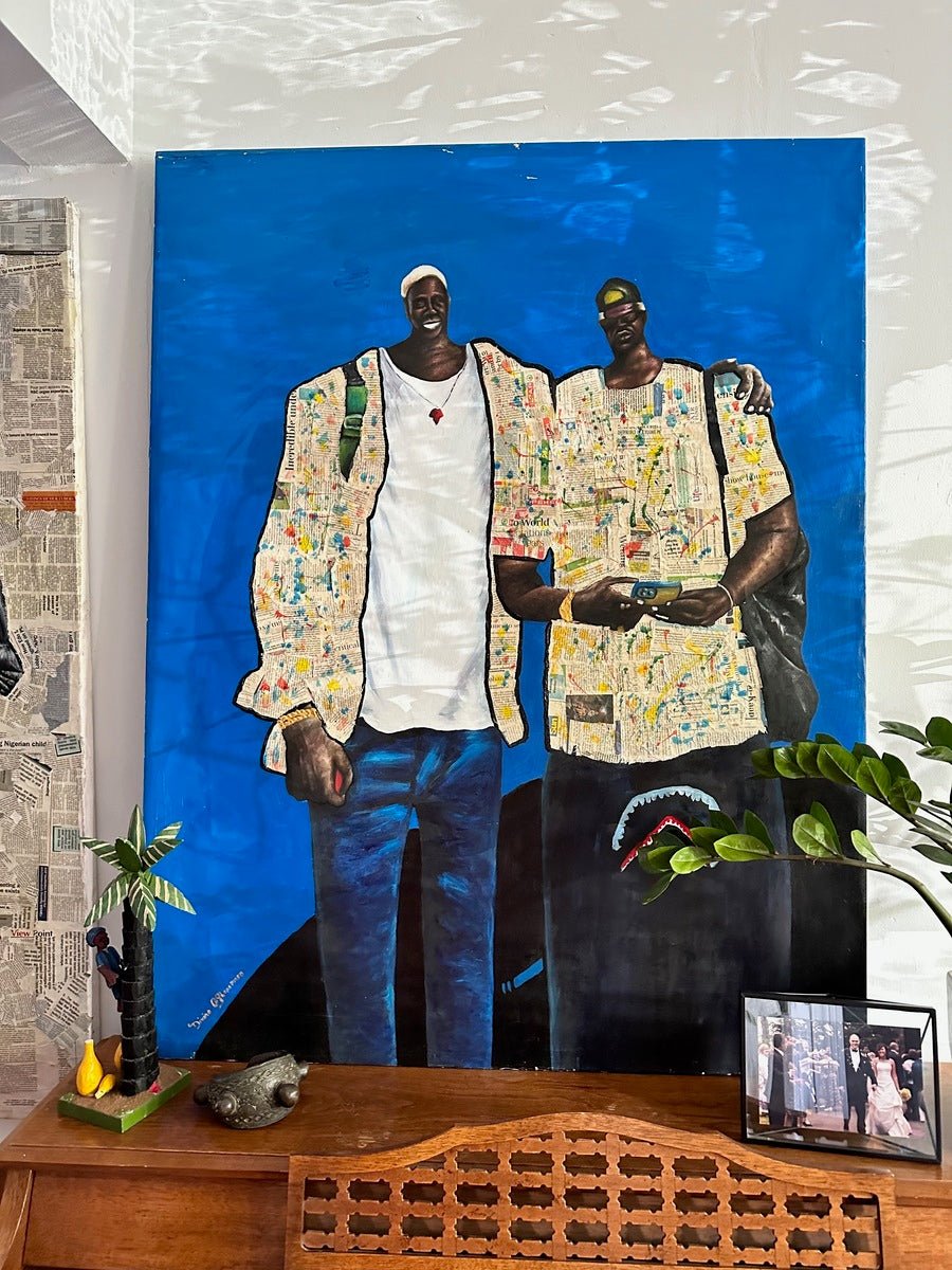 Friendship," a large hand-signed original painting by bibianna.co.uk, shows two casually dressed men side by side against a blue background. It hangs above a wooden table with plants, a decorative crocodile, and a framed photo.