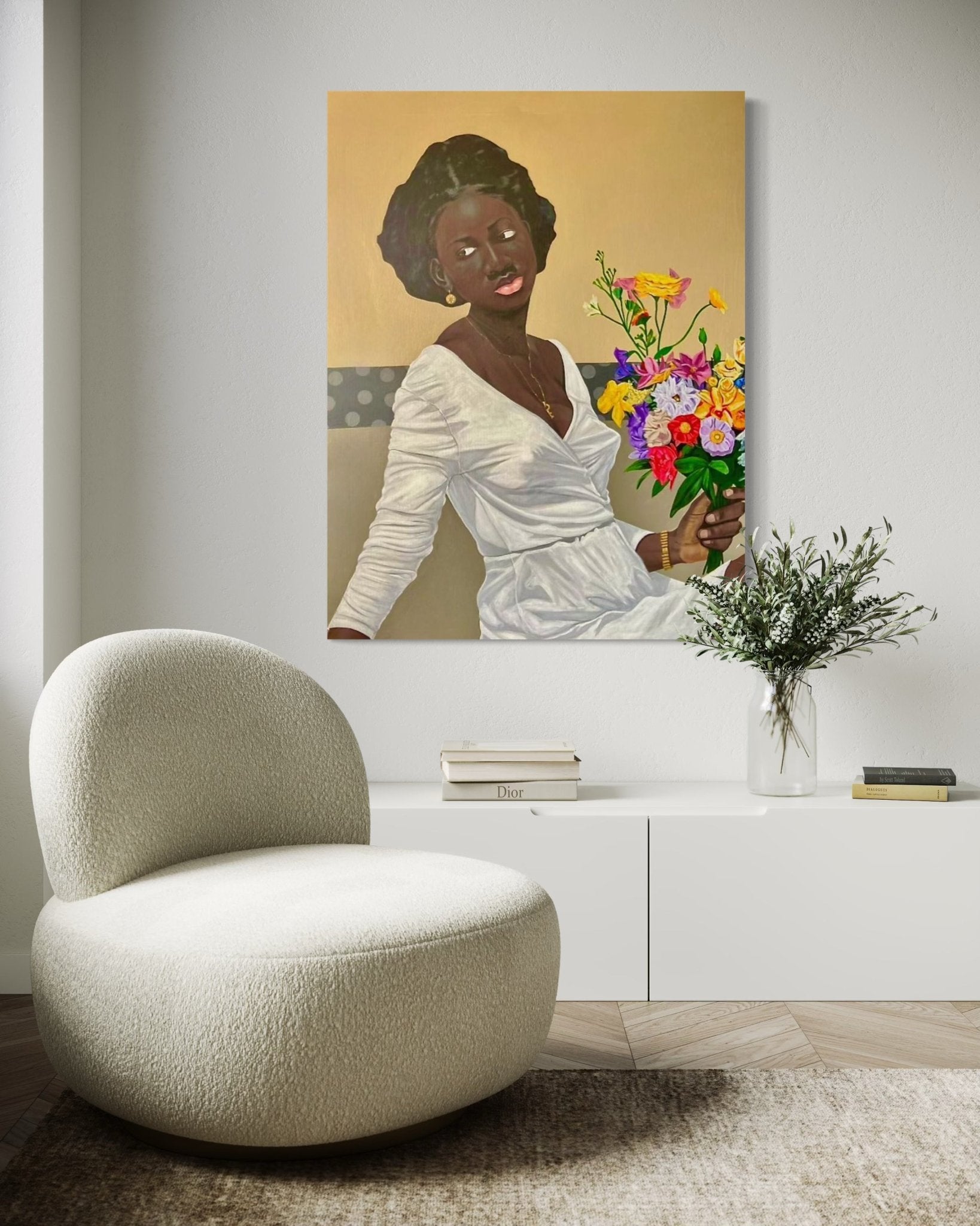 A modern living room featuring a rounded cream chair, a white console table with a vase of greenery, and "Give yourself all the flowers" by Ajenifuja Abiodun—an acrylic portrait of a smiling woman in white—hanging on the wall.