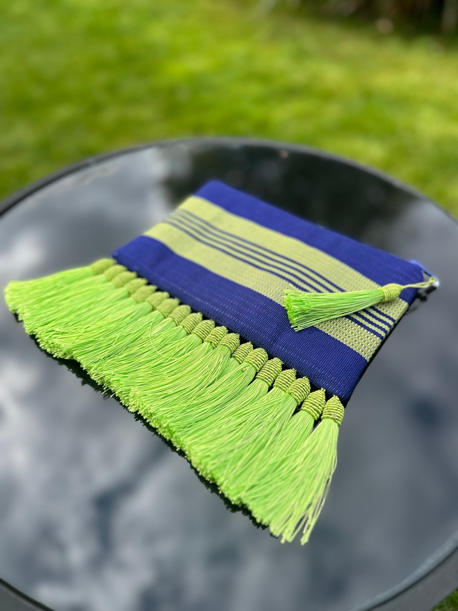 The Handmade Ashoke Tasseled Clutch by bibianna.co.uk features blue and green stripes with bright green tassels along one edge, displayed on a reflective black surface outdoors against a grassy background.