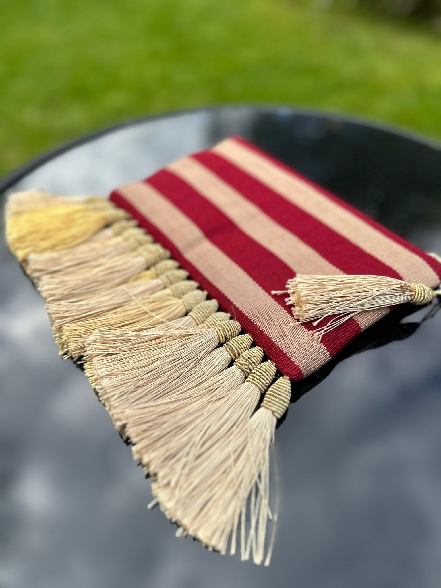 The Handmade Ashoke Tasseled Clutch by bibianna.co.uk, featuring red and beige stripes with long cream tassels, lies on a reflective black surface against a blurred green grassy background.