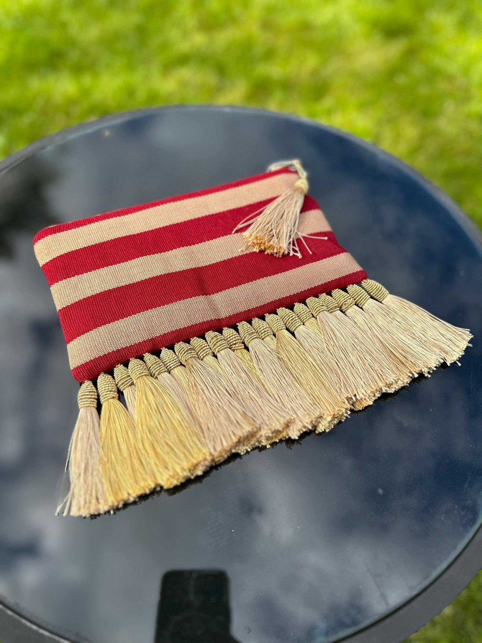 The Handmade Ashoke Tasseled Clutch by bibianna.co.uk features red and beige stripes, a zipper, and yellow tassels. Pictured on a shiny black surface with blurred green grass, it’s the perfect stylish occasion bag.