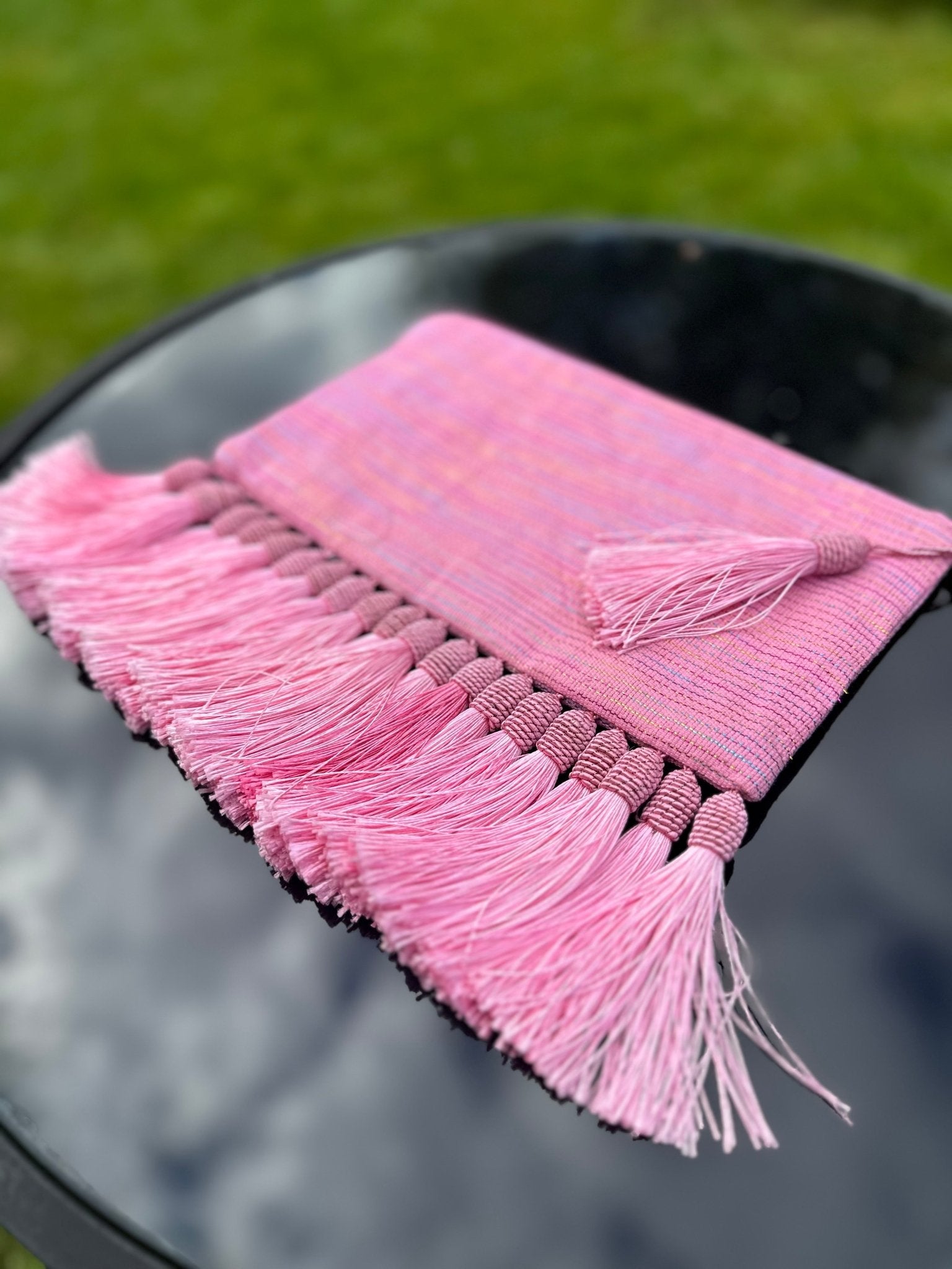 The Handmade Ashoke Tasseled Clutch by bibianna.co.uk features long pink and purple tassels with a matching zipper, shown on a round black table outdoors—perfect as a stylish occasion bag with grass in the background.