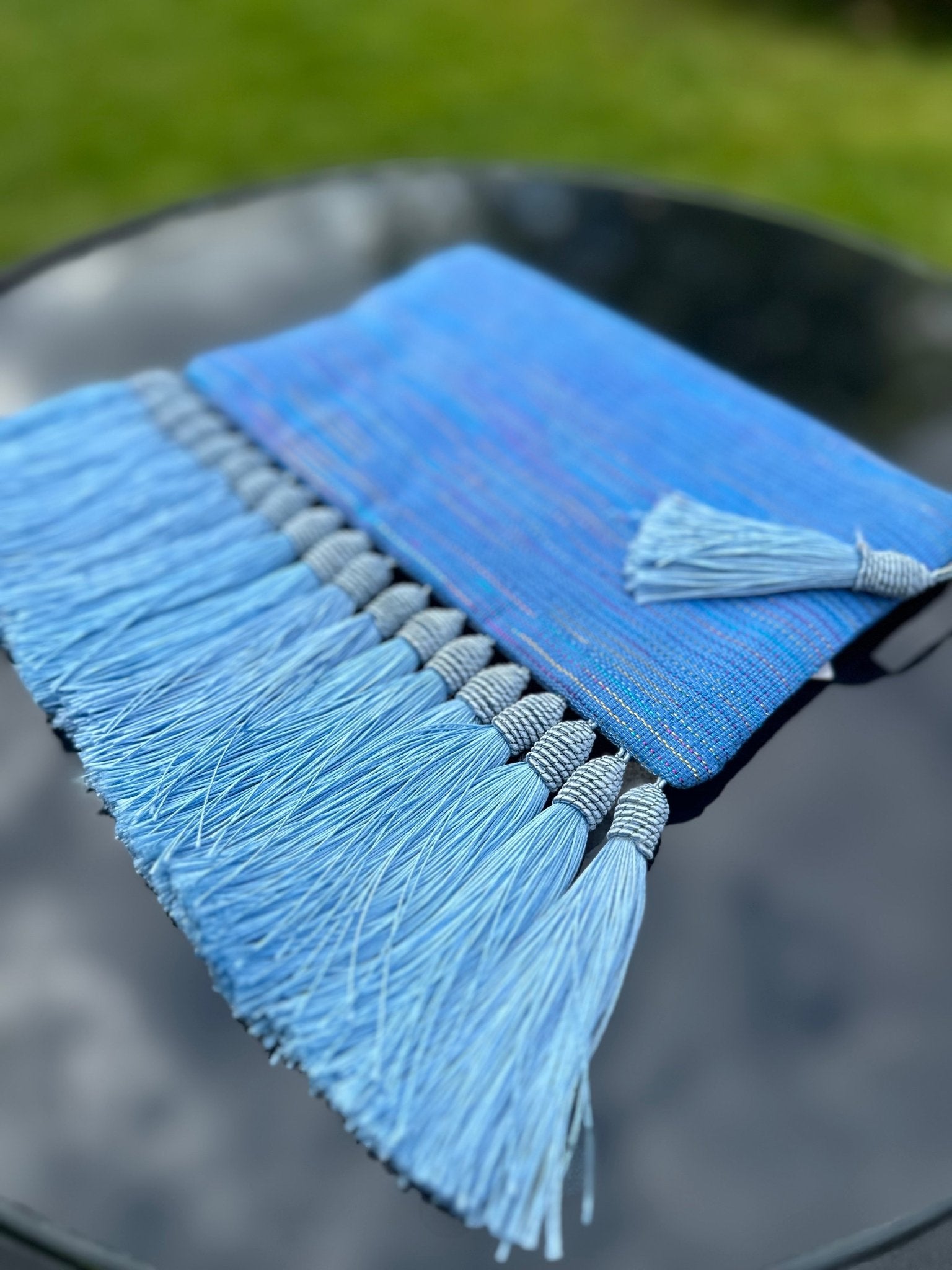 The Handmade Ashoke Tasseled Clutch by bibianna.co.uk—featuring a blue woven finish, matching tassel, and light blue fringe—rests on outdoor glass, green grass behind. The ideal stylish occasion bag for standout evenings.