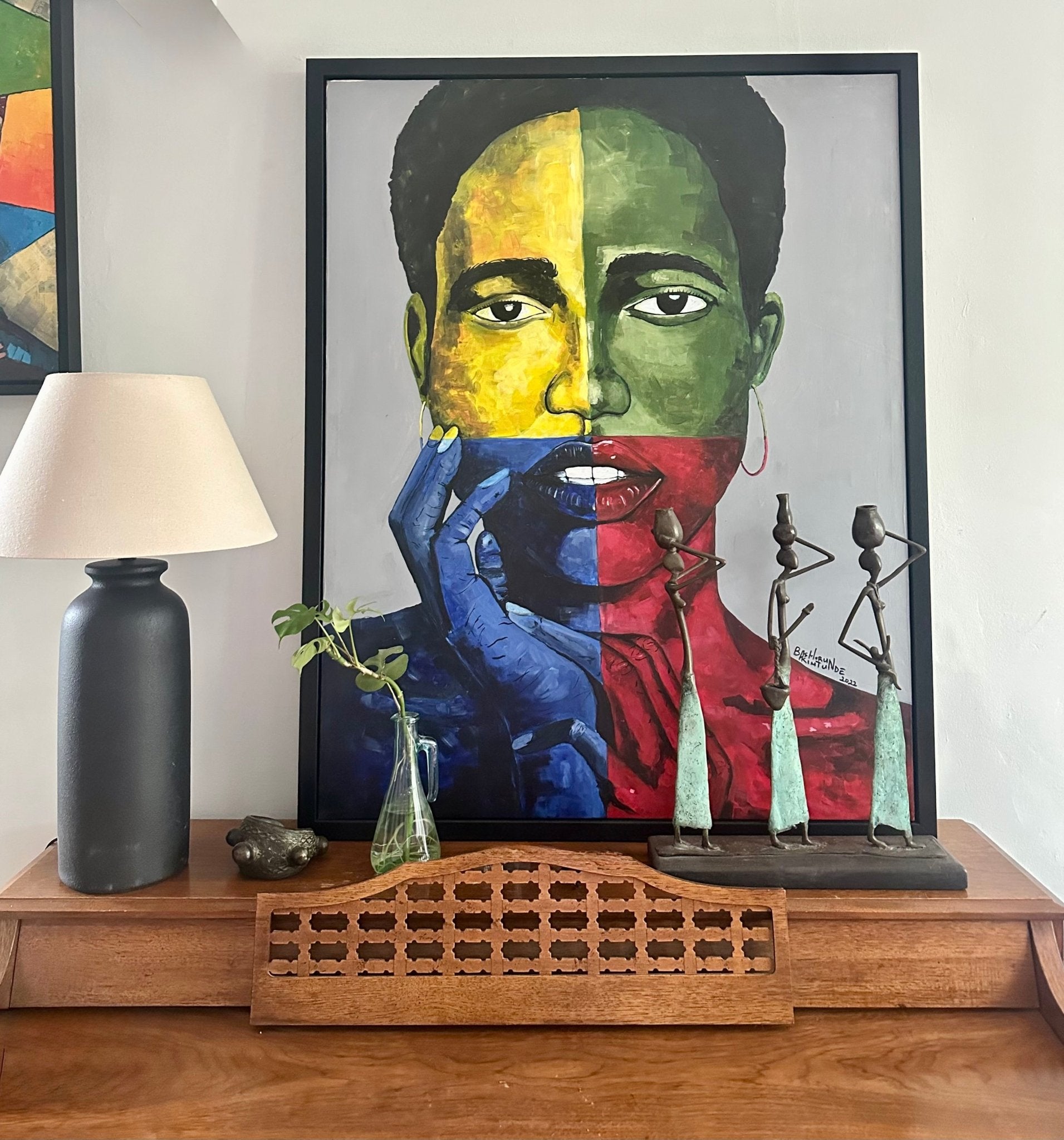 I Am Strong" Original Portrait Painting by Bashoru Akintunde, hand-signed and framed, features a face painted in red, yellow, blue, and green. It’s displayed on a wooden table with a lamp, glass vase plant, and three slender metal figurines.