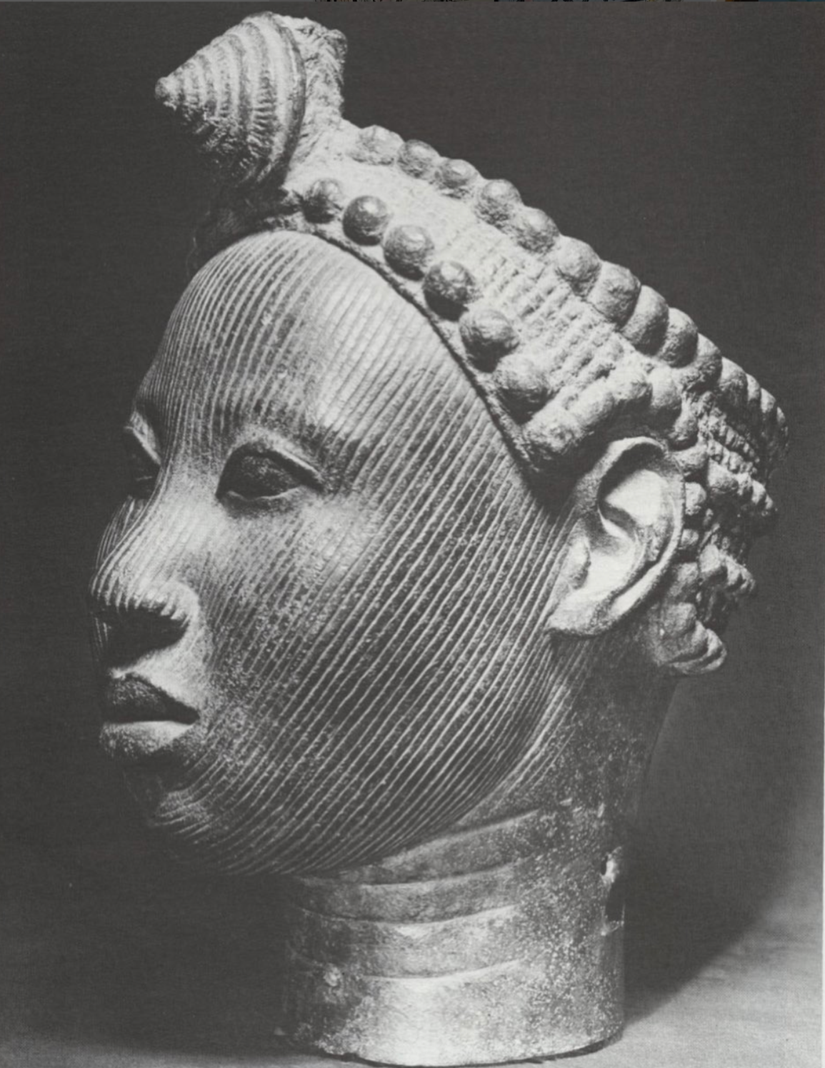 Black and white photo of a sculpted head with intricate facial markings, elaborate hairstyle with beads or knots, and a calm, composed expression. The sculpture appears to be made of bronze or metal.