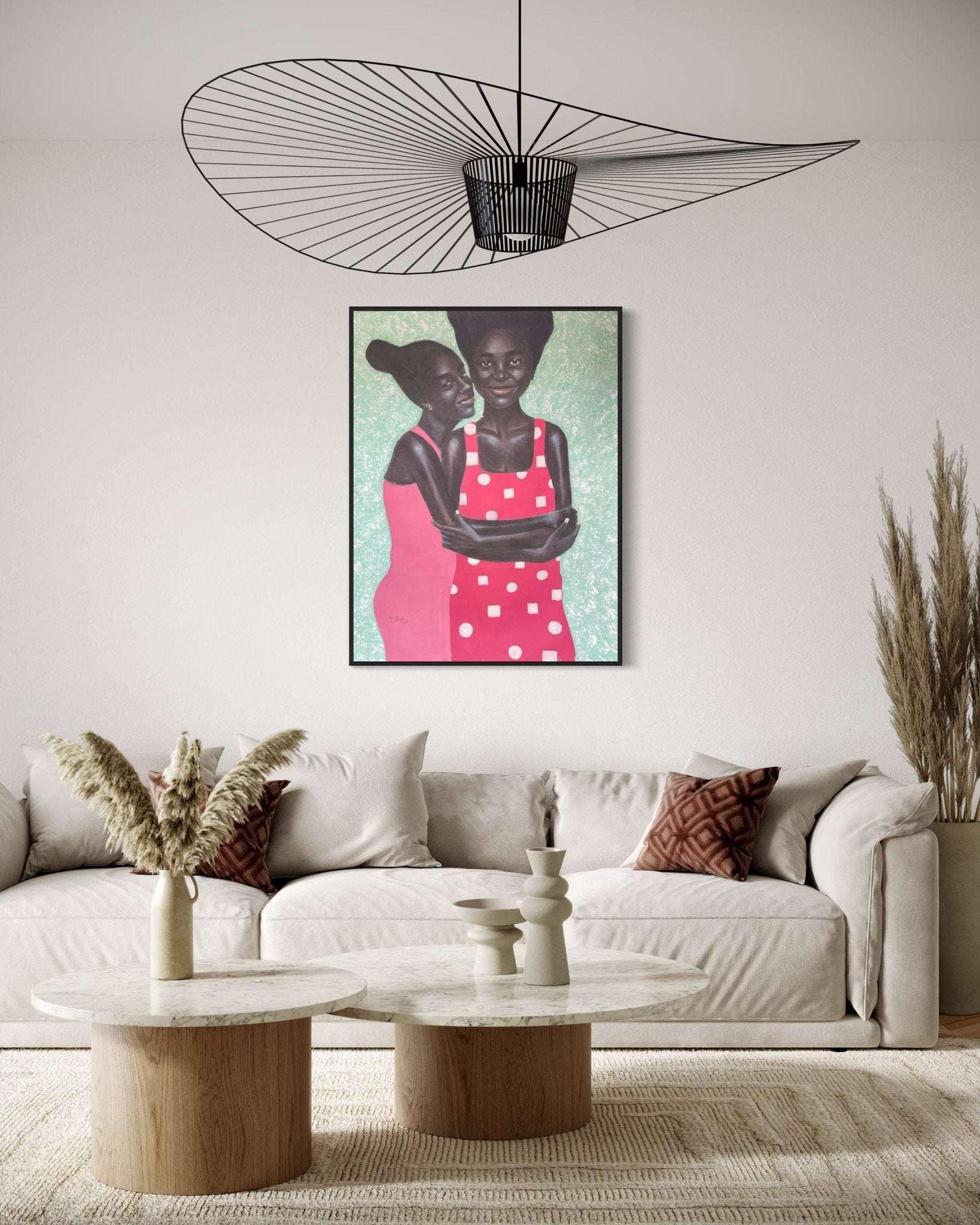 A modern living room with a beige sofa, two round coffee tables, vases, pampas grass, a wire pendant light, and "Joyful African Sisterhood Portrait in Vibrant Colors, 'Sisterhood'" wall art by Henry James.