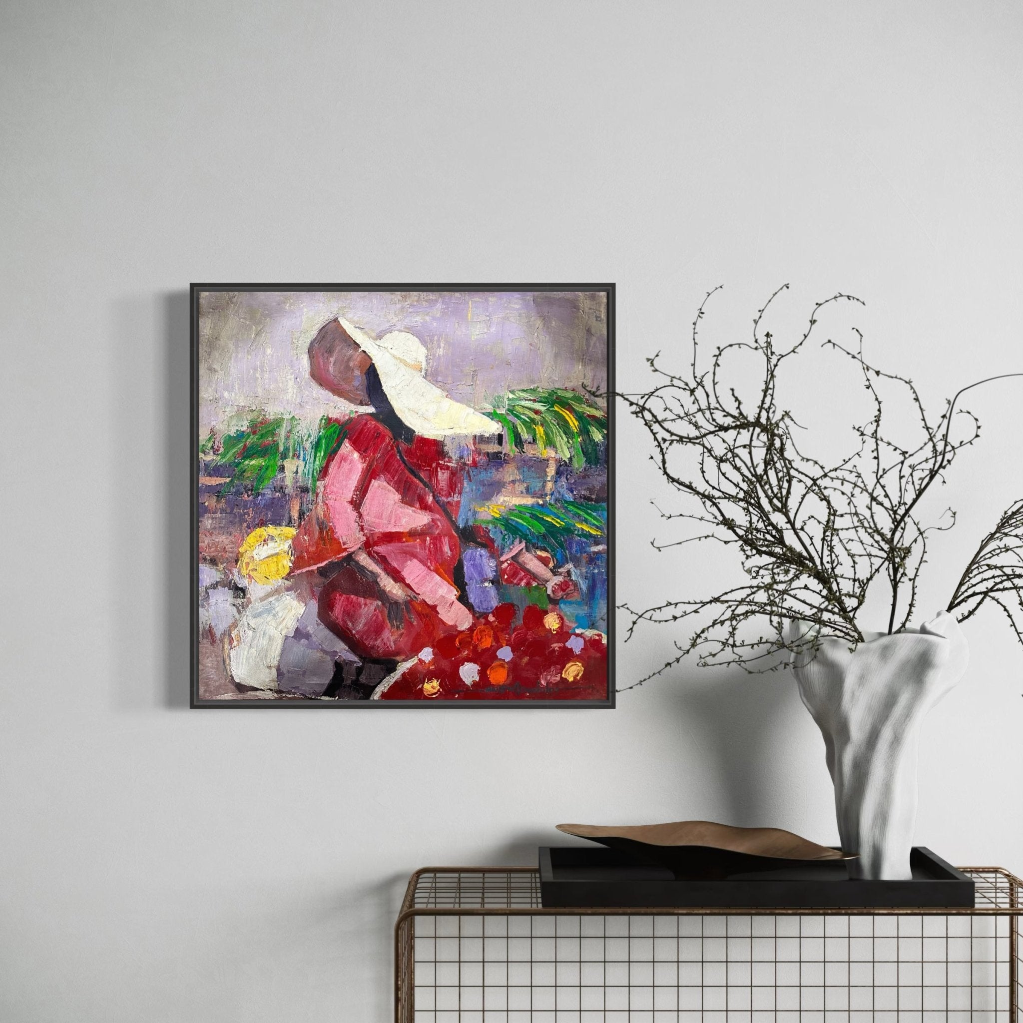 The "Market Sance" Lady In Sun Hat framed painting by NOSA OSADOLOR shows a woman in a pink outfit and sun hat among red flowers, displayed on a white wall above a wire console table with a white vase of bare branches.