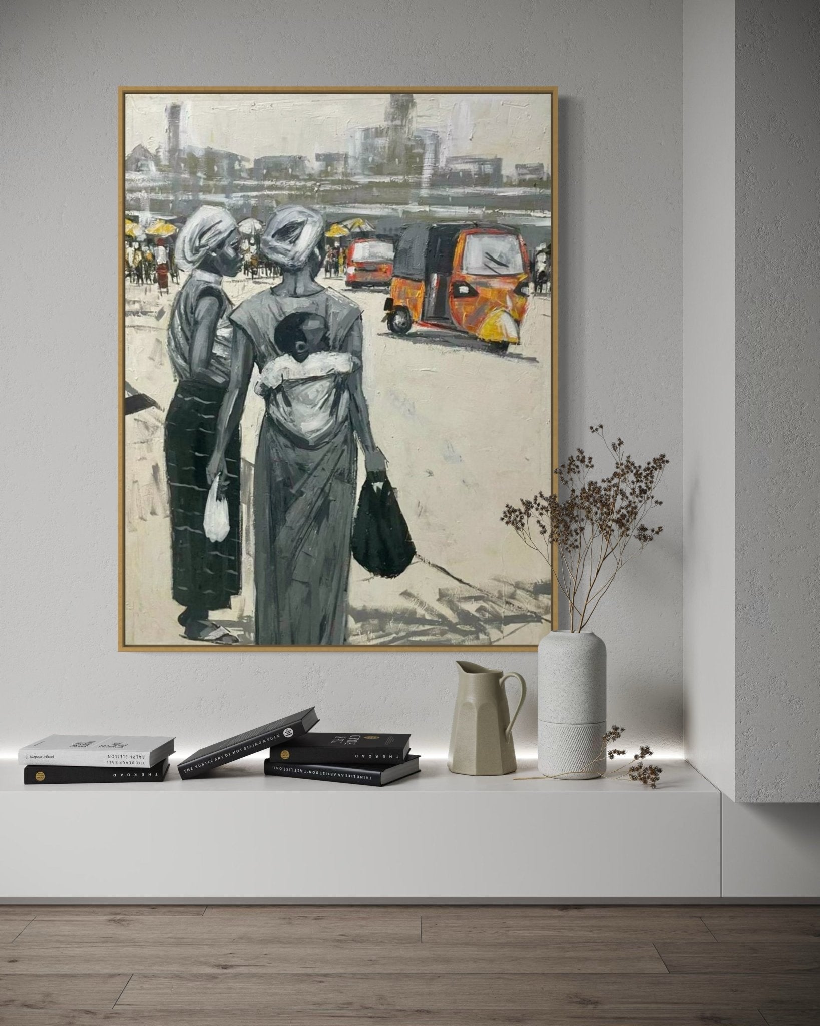 'Lagos Life' City Scape Still Life by Lucky Isaiah features two women in traditional attire with a baby beside bright auto rickshaws, framed and displayed above a white shelf with books, a vase, and dried flowers in a modern room.