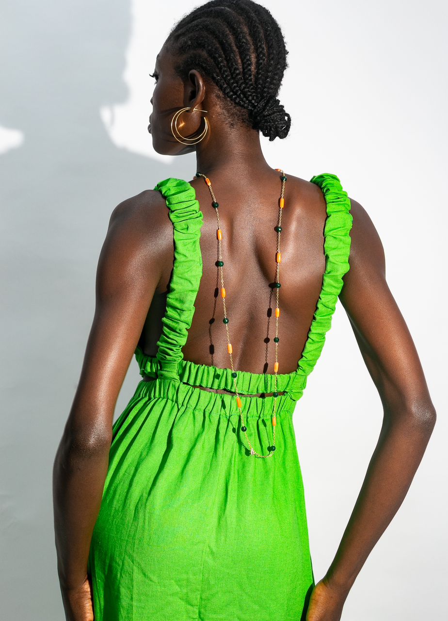 A person with braided hair and gold hoops wears a bright green Linen Cutout Dress by bibianna.co.uk, featuring a low back and ruffled straps, styled with a long beaded necklace draped down their back.