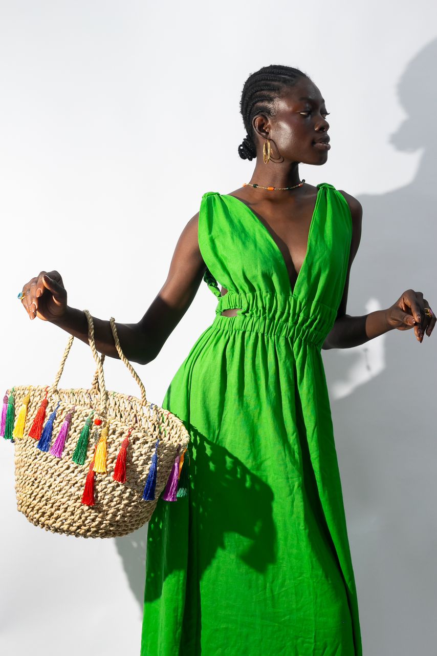 A woman in braids models the Linen Cutout Dress by bibianna.co.uk, paired with a straw tote bag adorned with colorful tassels. She poses against a white background, gazing to the side in this stylish summer look.