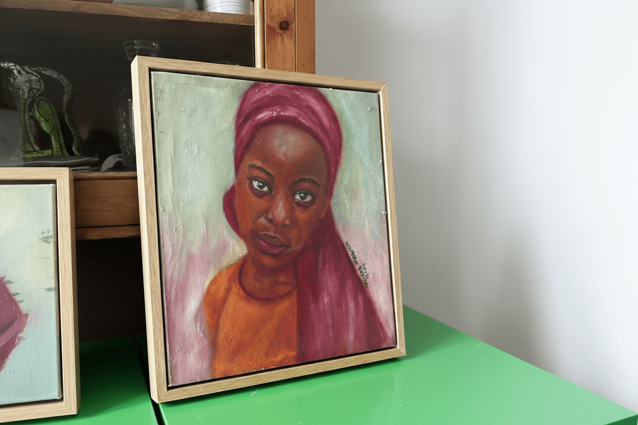 The "Little Amina" framed portrait by Bibianna African Art Collective features a woman in a pink headscarf and orange top on a green surface, beside wooden shelves with glassware, gazing calmly against a soft background.