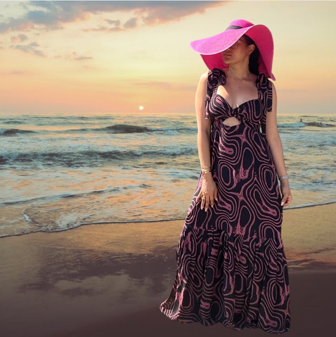 A woman in the Magnolia Maxi Dress by bibianna.co.uk and a wide-brimmed pink hat stands on a beach at sunset, with gentle waves and an orange-pink sky in the background.