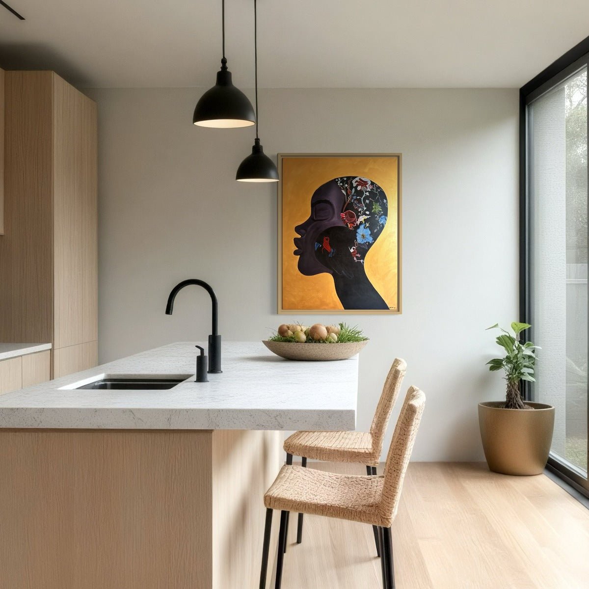 Modern kitchen with light wood cabinets, white island with black faucet, two woven chairs, bowl of fruit, pendant lights, potted plant, and "Manifesting" profile portrait artwork by Azizo on the wall.