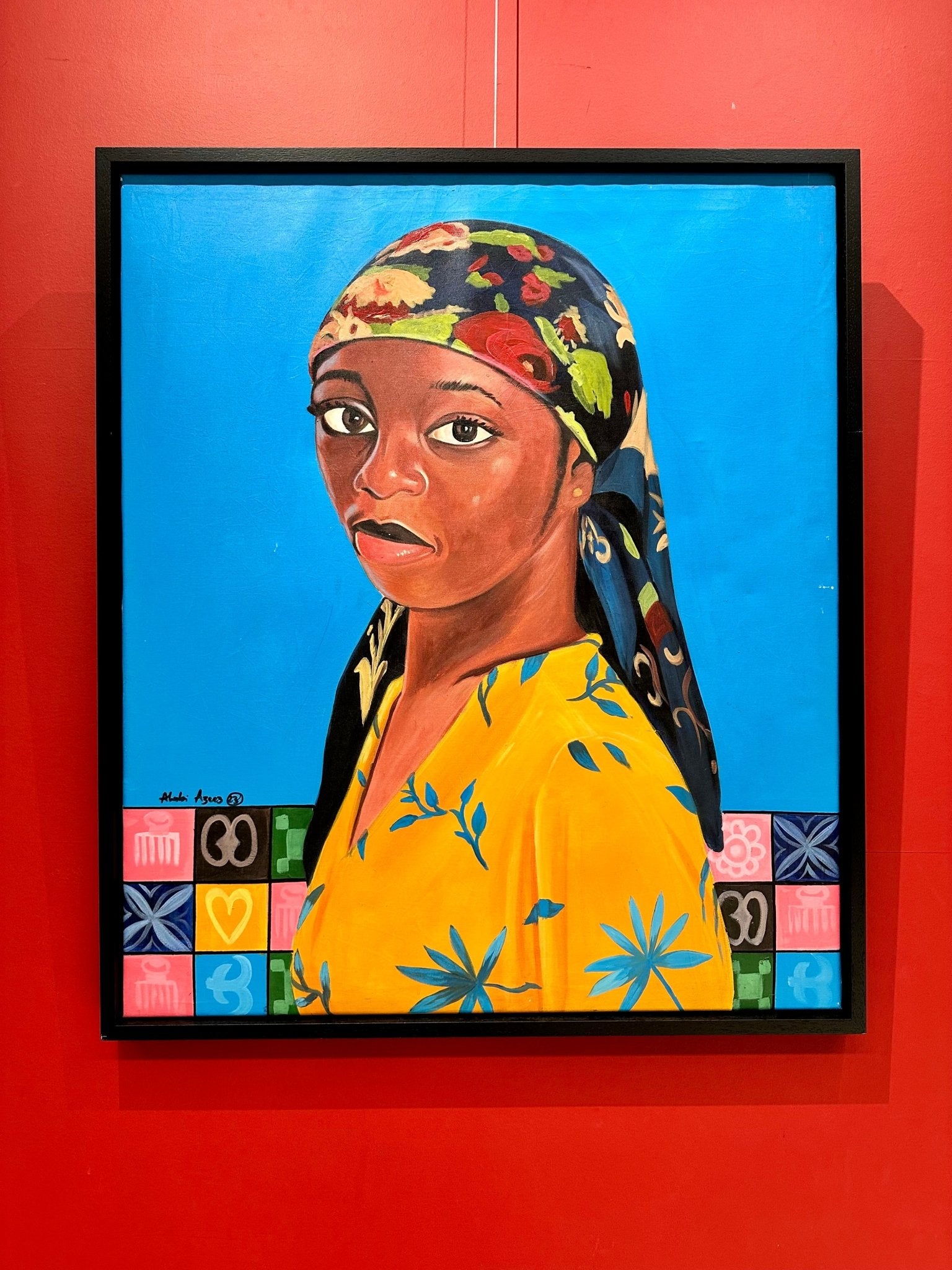 Marthas Flowers" Portrait by Alabi Azeez: a framed acrylic on canvas of a young Black woman in a yellow floral dress and vibrant headscarf, set against a bright blue background with multicolored symbols, displayed on a red wall.