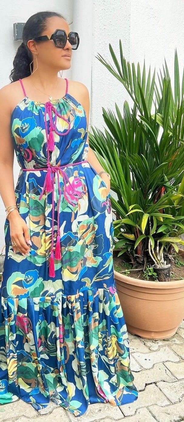 A woman in sunglasses and a vibrant Meena Dress from bibianna.co.uk stands beside a potted green plant outdoors, with one hand in her pocket and the other by her side.