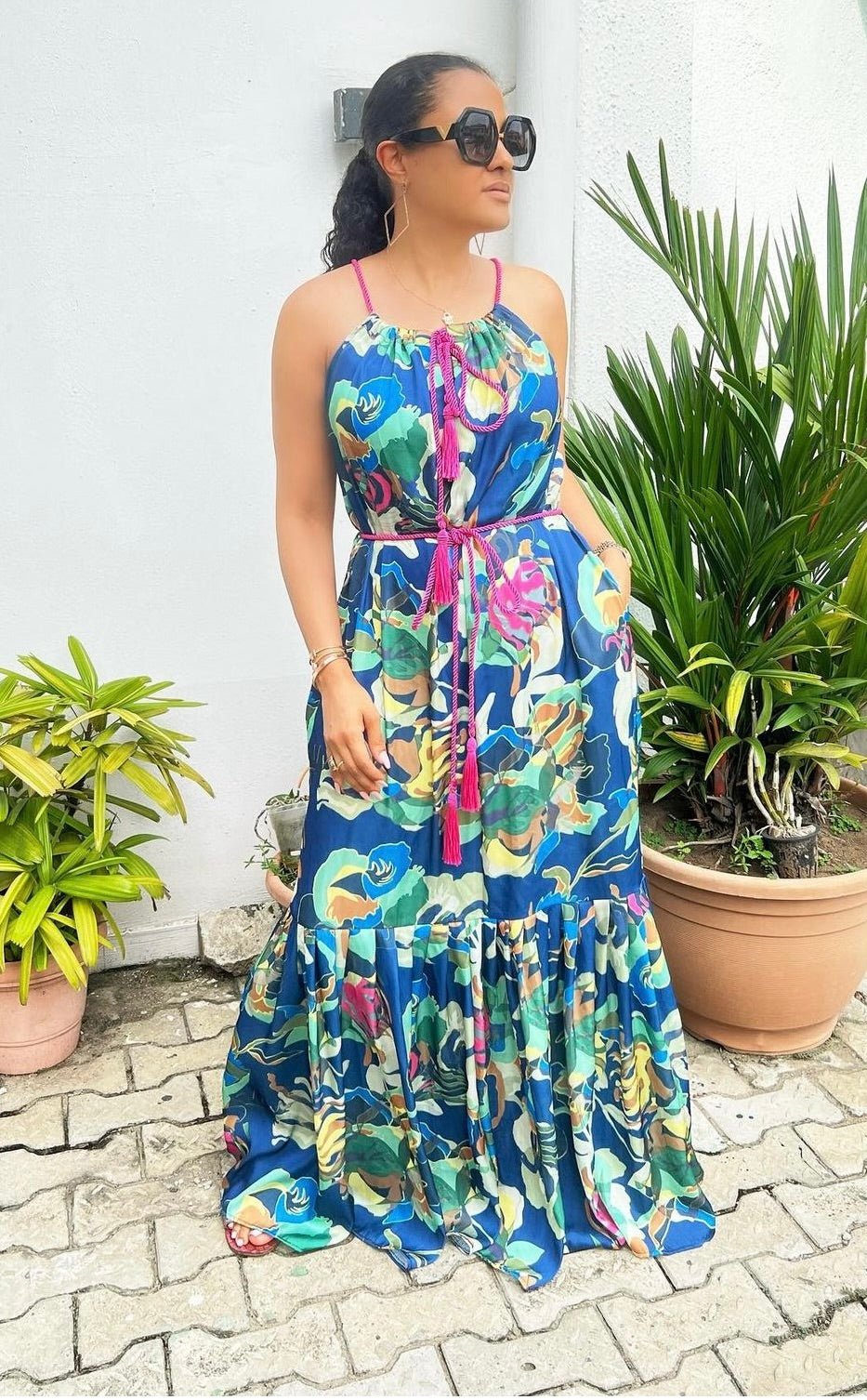 A woman in the Meena Dress by bibianna.co.uk, with a vibrant floral print and oversized sunglasses, stands on a paved area beside potted greenery, looking to the side with one hand in her pocket.
