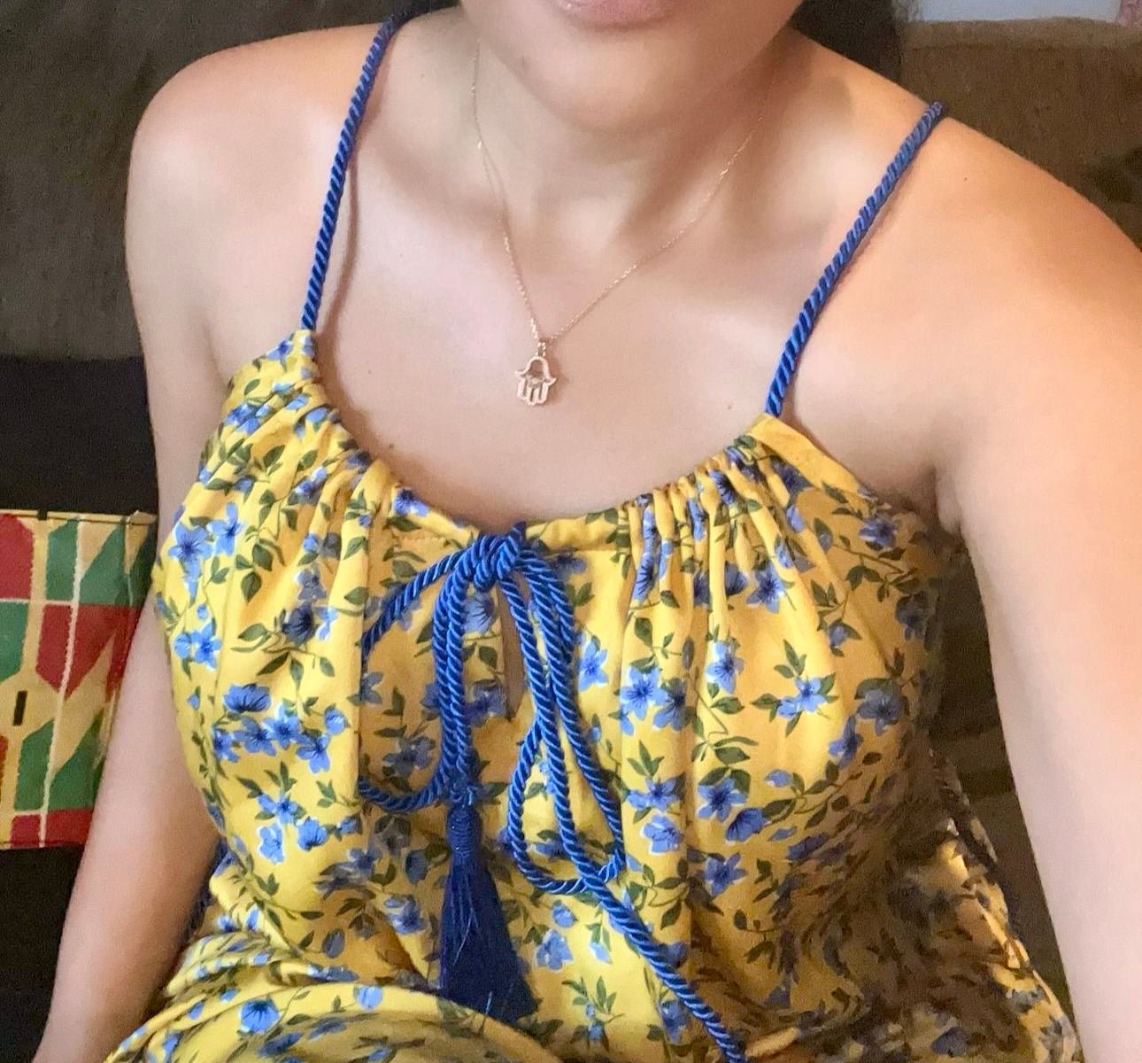 Someone wearing the Meena Dress by bibianna.co.uk—a yellow, blue floral print dress with blue straps—is seated on a couch. A small charm necklace adorns their neck, and their face is partially out of frame.