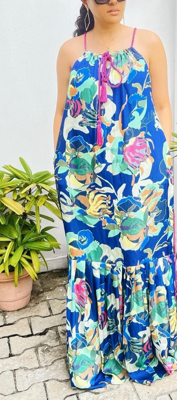 A woman in the Meena Dress by bibianna.co.uk—a long, colorful floral print dress with pink straps and tassels—stands outdoors on a stone path beside a green potted plant, wearing sunglasses with her face partially out of frame.