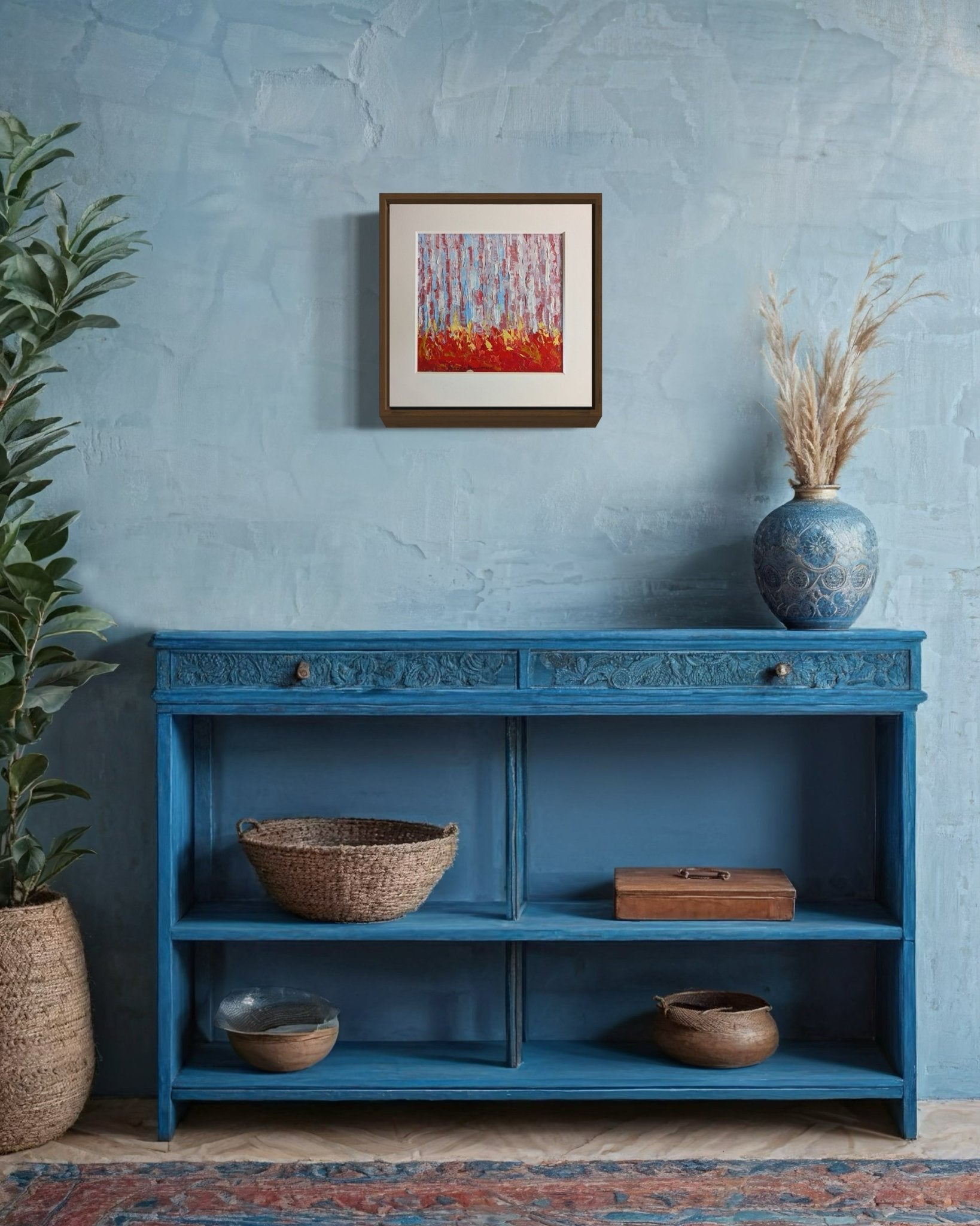A blue console table with baskets and a box sits against a textured light blue wall. Above, Miniature Abstract Landscape Kehinde Badmus by Kehinde Badmuss adds flair, alongside a vase with dried grasses and a leafy plant.