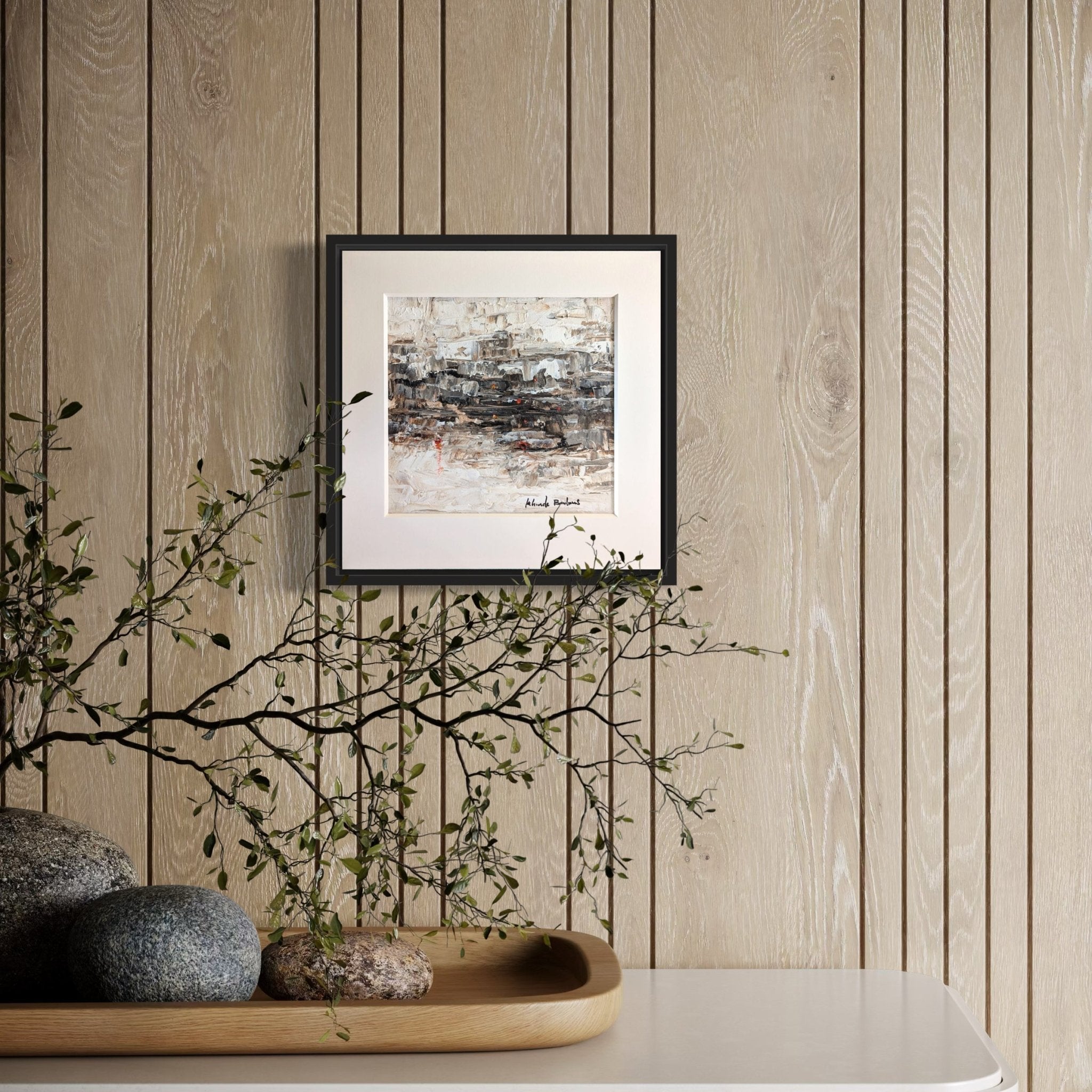The Miniature Abstract Landscape by Kehinde Badmus, framed and painted in neutral acrylics, hangs on a light wood wall. Below it, a tray with smooth stones and a branch complements the minimalist look.