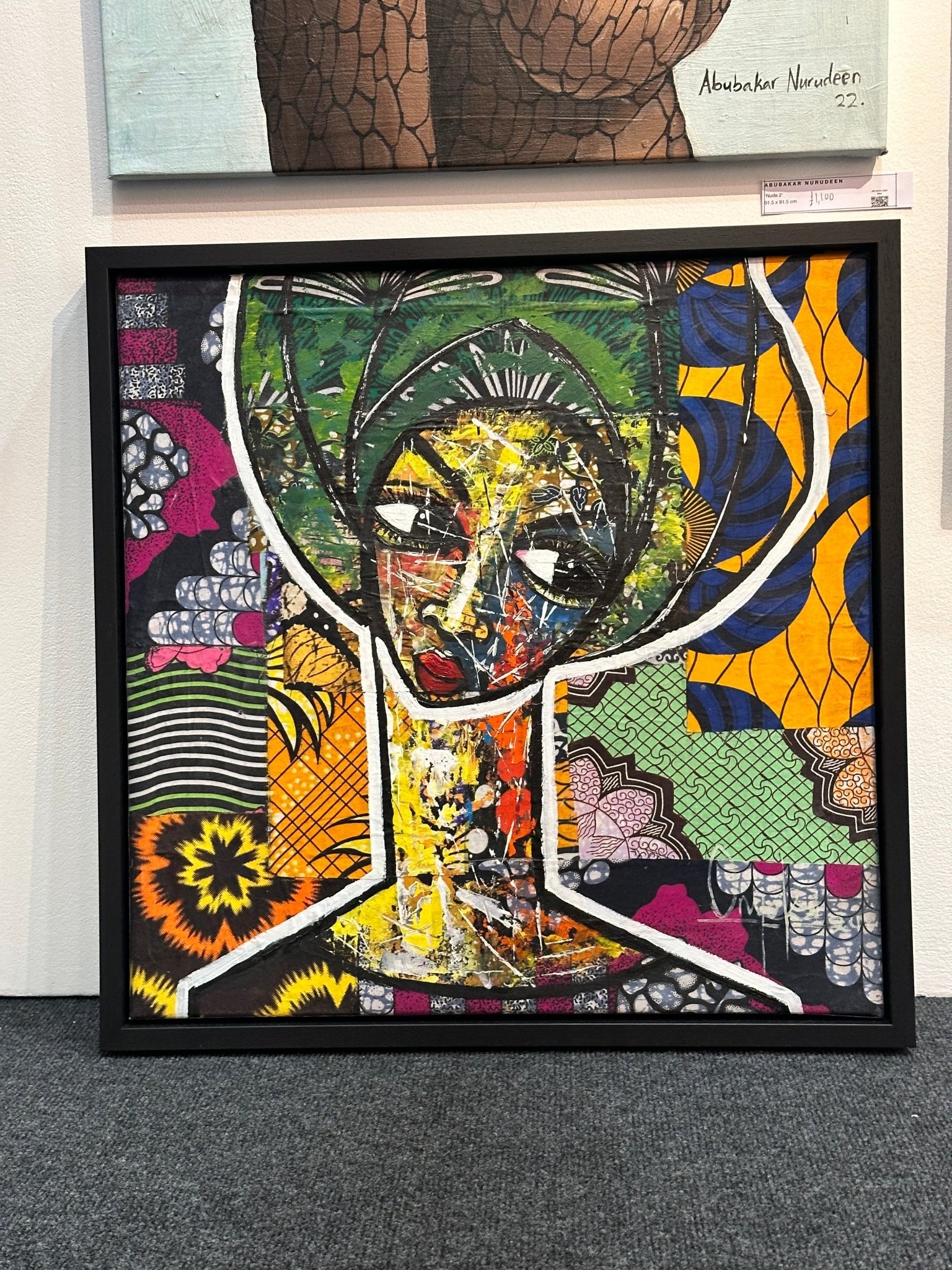 Maiden" by Onyeka O is a framed mixed media collage using Ankara fabric and acrylic, depicting a stylized figure with bold lines and vivid colors, displayed on a carpeted floor against a wall.