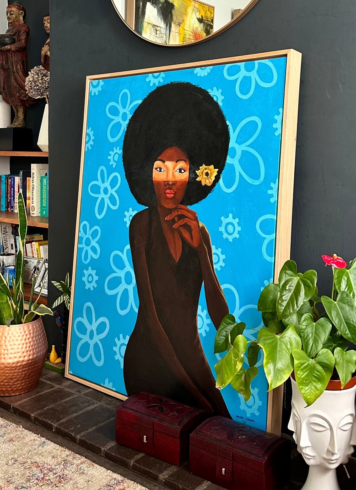 The framed acrylic painting 'My Afro 2' by Opeyemi (Afuope Opeyemi) features a woman with an afro and yellow flower amid blue floral motifs, displayed on a hearth alongside plants and books in a modern, stylish room.