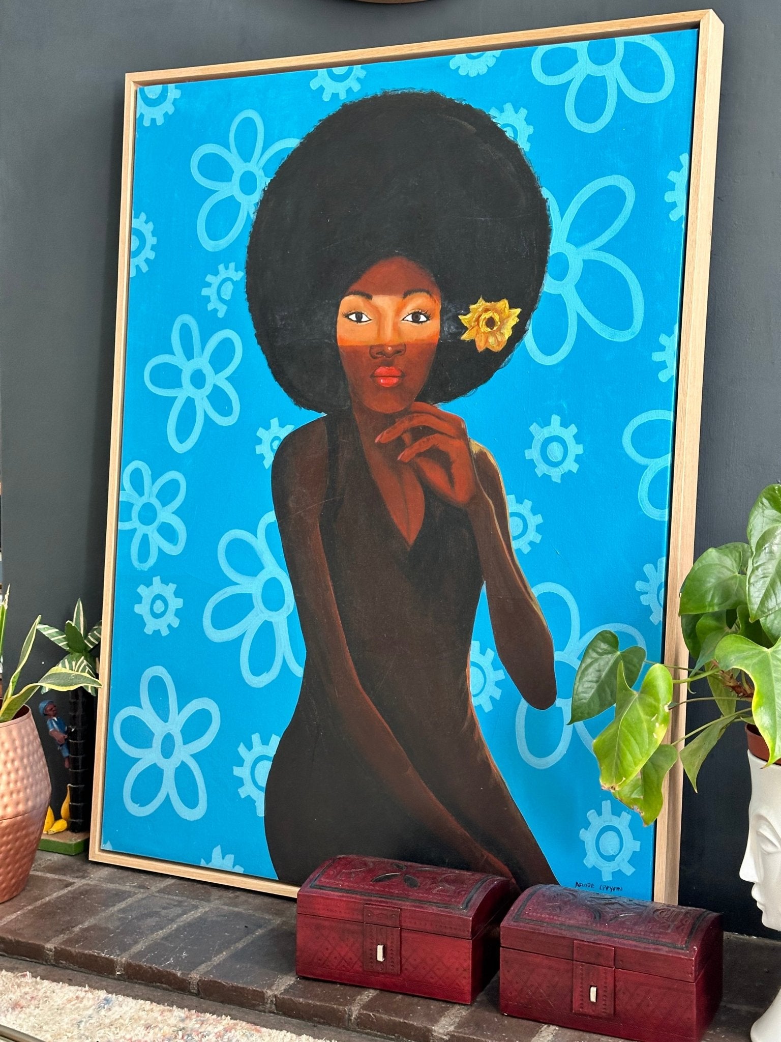 'My Afro 2' by Afuope Opeyemi is a framed acrylic on canvas featuring a woman with an afro and yellow flower, set against a blue and white floral background. The retro style artwork is displayed near two red boxes and a green potted plant.