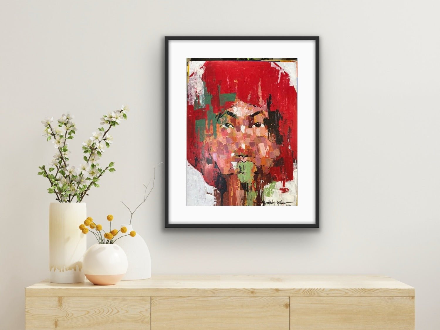 A framed "One of a Kind Series by Oyelusi" original by Olasunkanmi Oyelusi, featuring bold red, green, and orange acrylic on paper, hangs above a light wood cabinet with a white vase of flowers and a yellow-accented ceramic bowl.