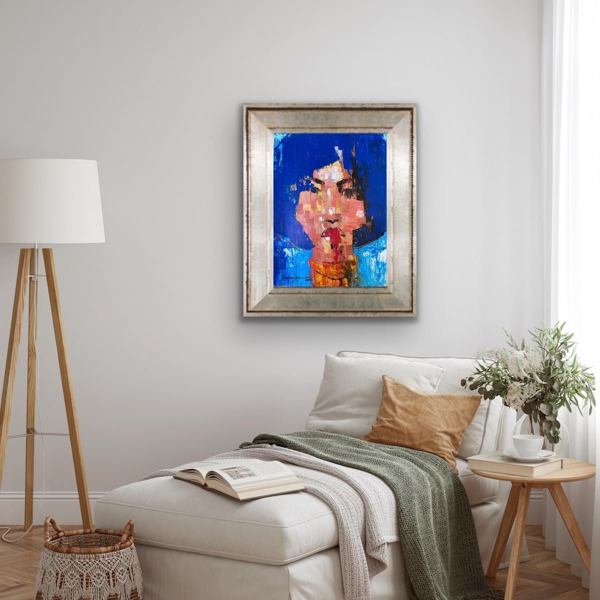 A cozy living room features a white armchair with a green throw and beige pillow. Above hangs "One of a Kind Series by Oyelusi" from Olasunkanmi Oyelusi, an acrylic painting in blue and pink, framed. A lamp, basket, book, and plant finish the look.