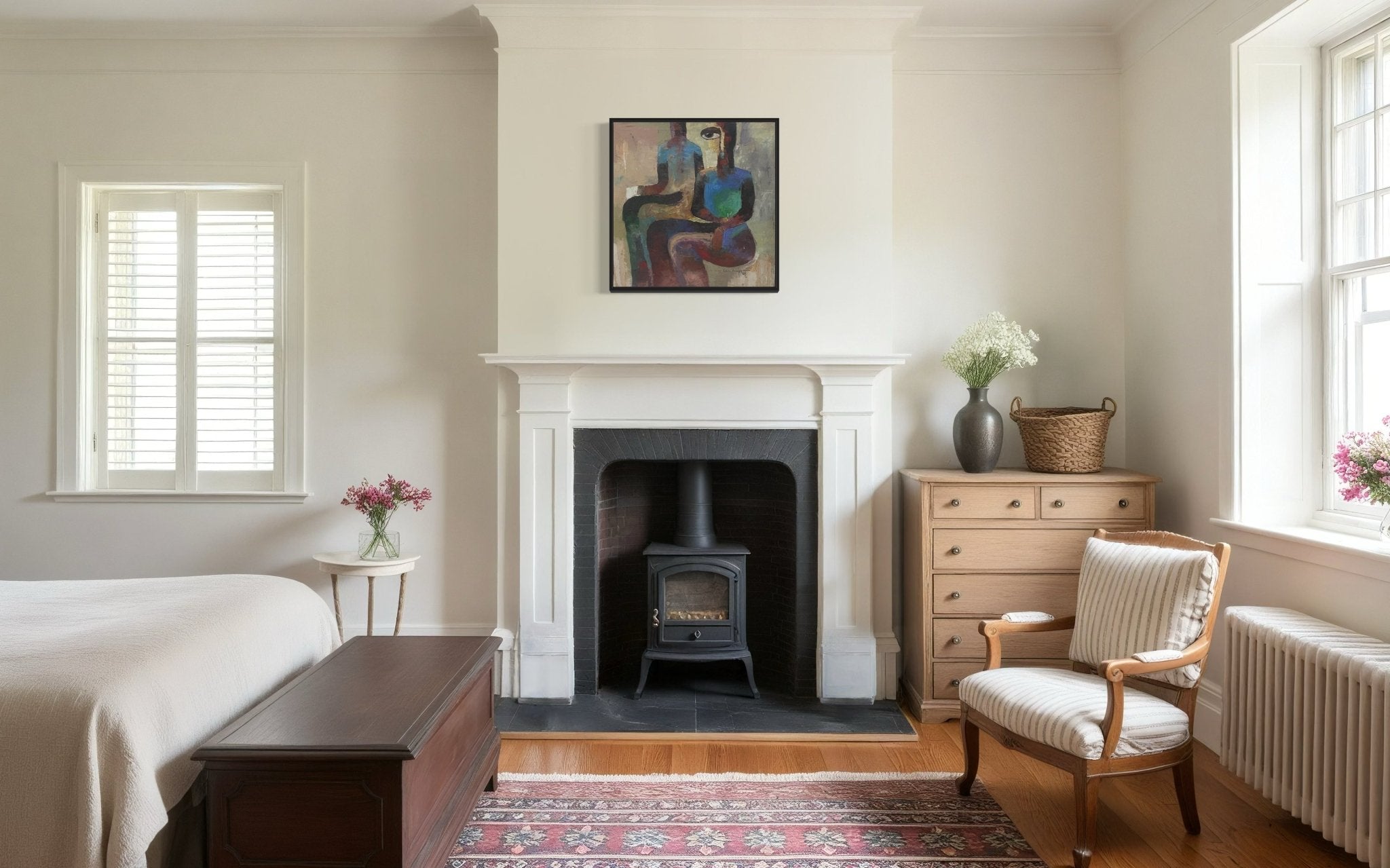 A cozy bedroom with light walls, a fireplace featuring a black wood stove, and a bed. Above the mantel hangs "Pension Cue" by Achike Anayo, hand-signed and including a certificate of authenticity. Sunlight streams through the windows.