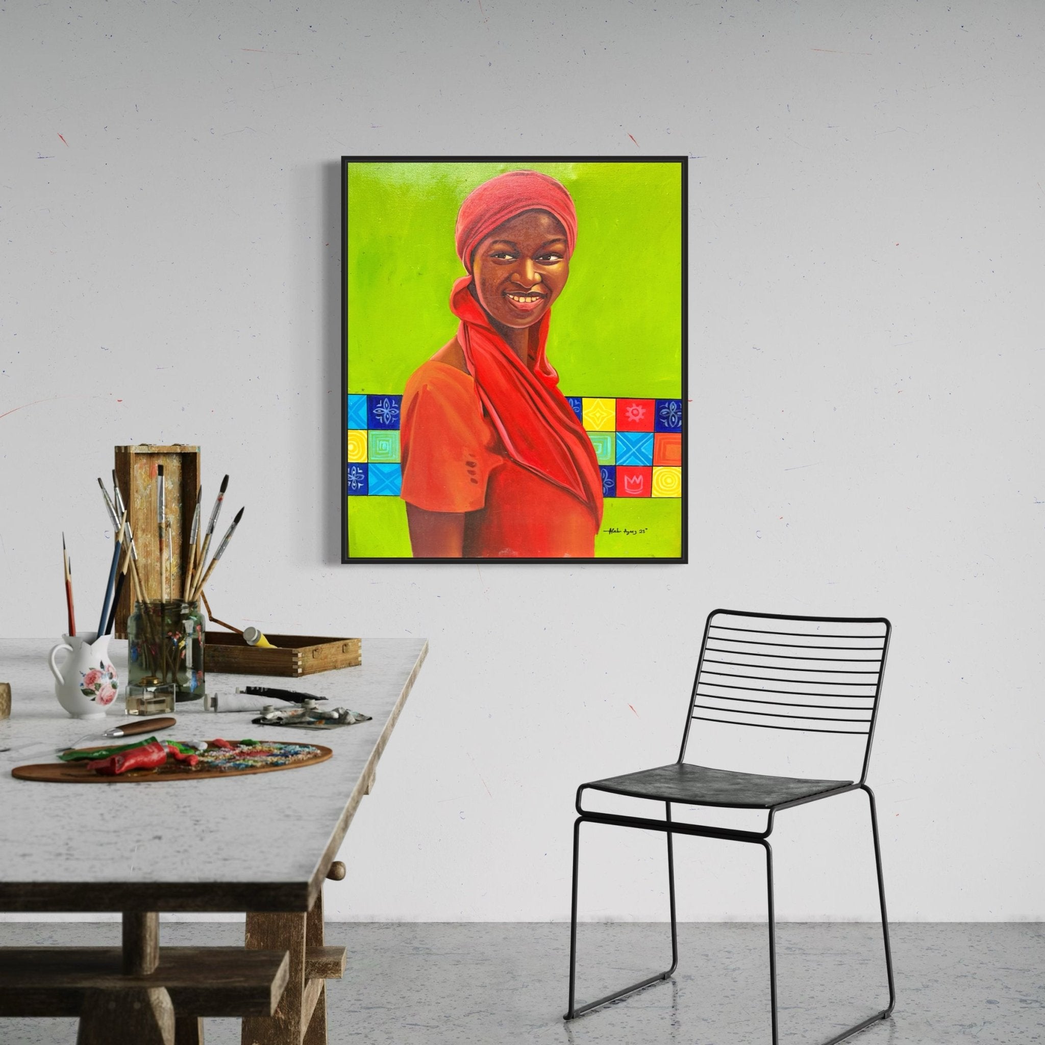 A modern art studio with a table, wire chair, and "Red Scarf" by Alabi Azeez—an acrylic on canvas of a smiling woman in red clothing and headscarf, set against a vivid green patterned background.