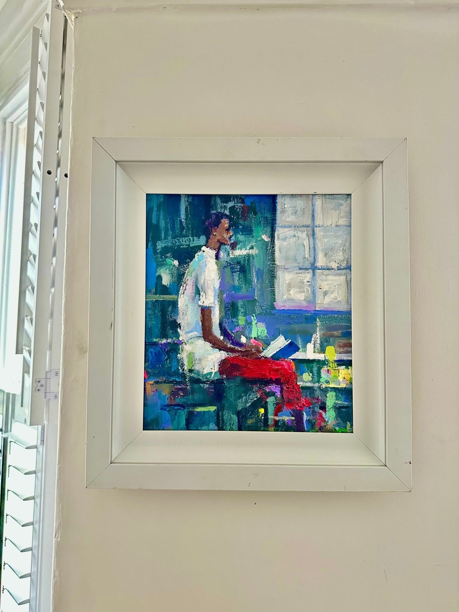 Waiting" by Wande George is a vibrant acrylic on canvas in a white frame, featuring a person in a white shirt and red pants reading sideways, set against abstract blue and green hues with light streaming through a window.