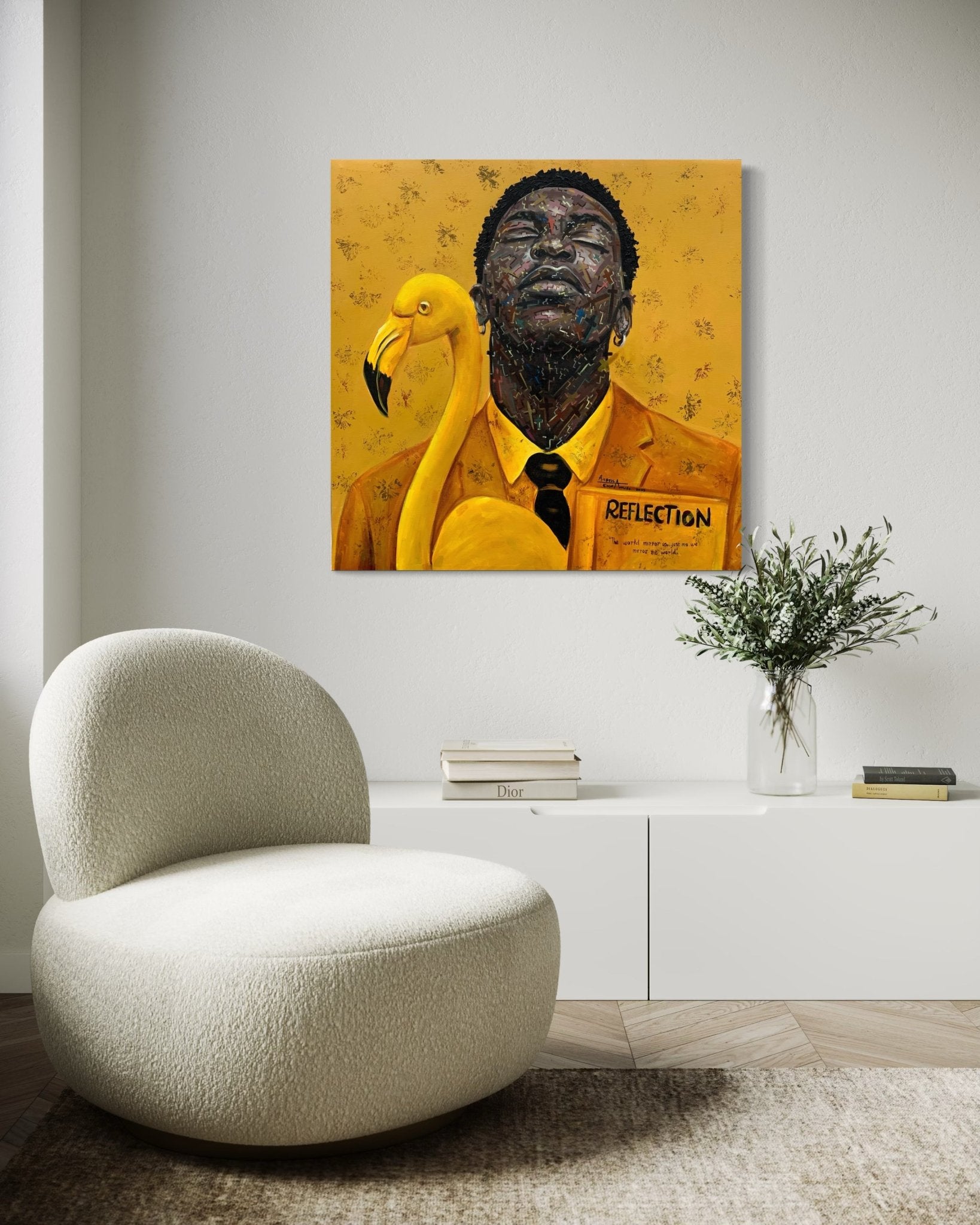 A modern living room with a curved white chair, low white cabinet, and greenery. On the wall is "Reflection," an original painting by Agboola Emmanuel I, showing a suited man and a flamingo from the Finding Self Series.