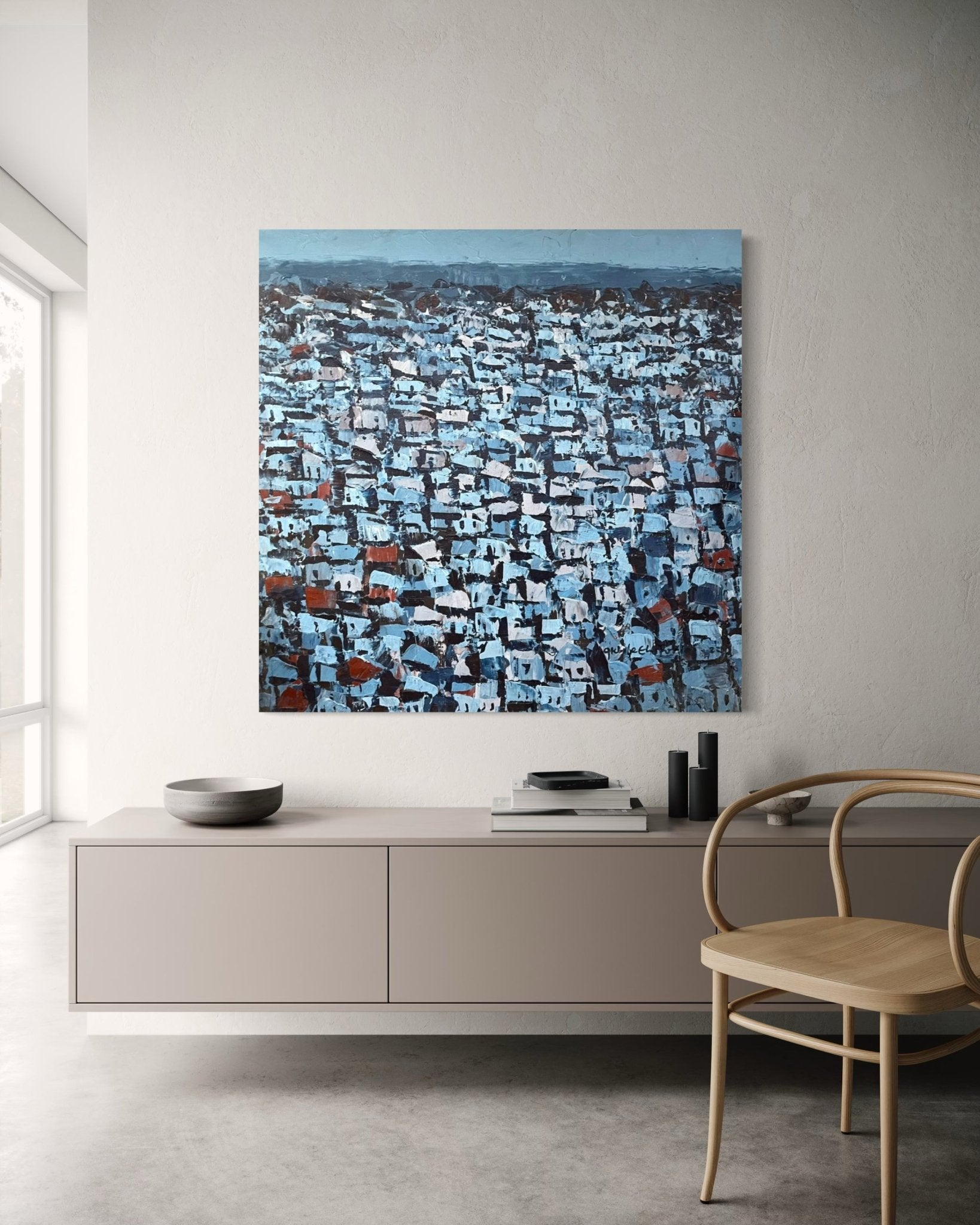 A modern, minimalist living room features light walls and floors, a wooden chair, a beige sideboard with decor, and "Rooftops Series," an acrylic on canvas cityscape in cool blue and gray by Akhile Ehiforia, hand-signed by the artist.