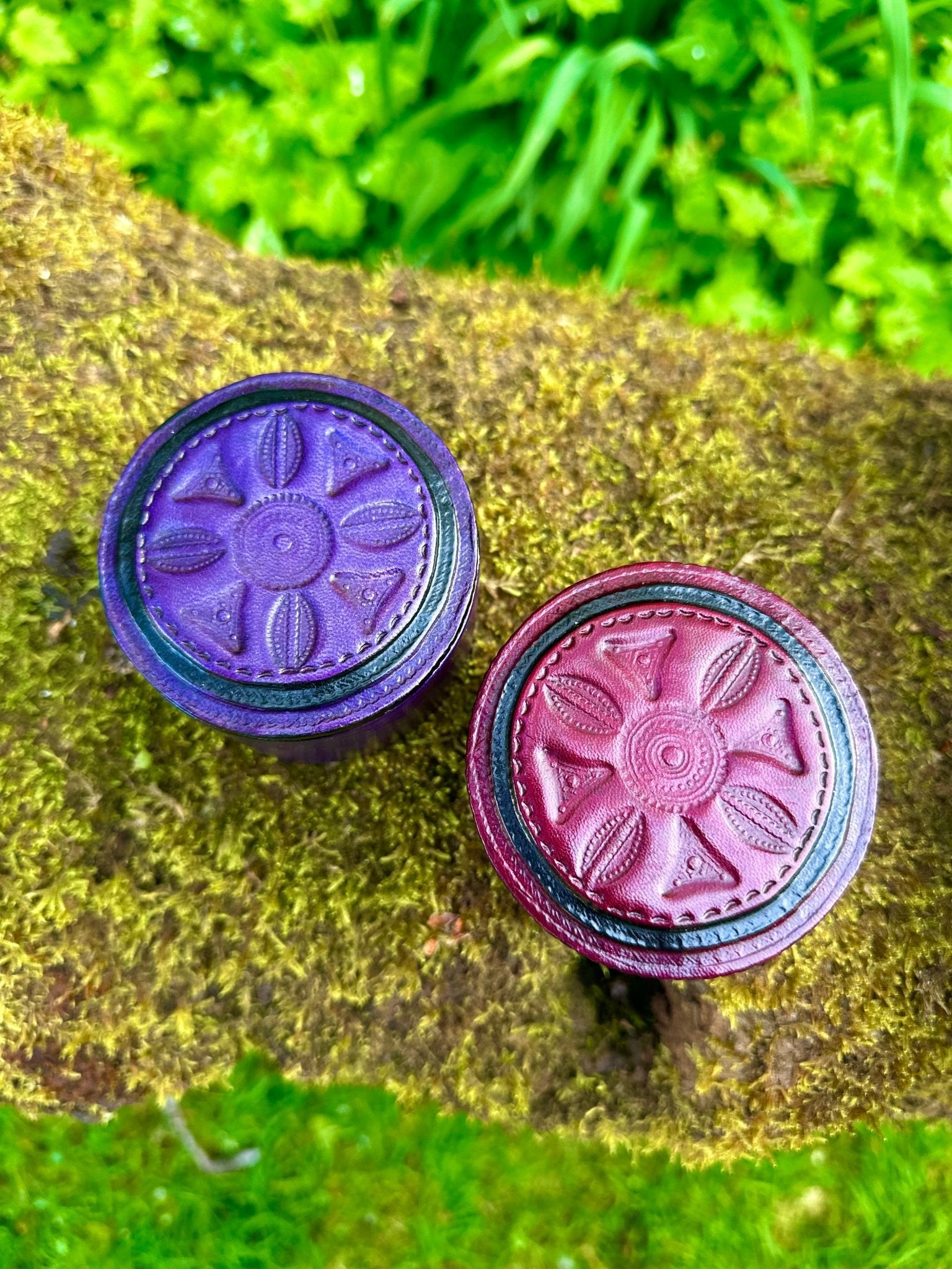 Two round bibianna.co.uk boxes—a purple African Tuareg hand-tooled leather trinket/jewellery box and a maroon embossed box—sit on mossy ground with green foliage behind.