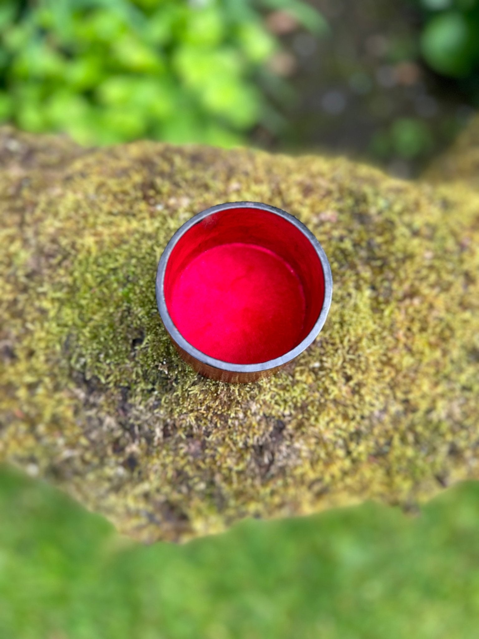 A round metal cup with a bright red interior rests on moss, next to the bibianna.co.uk Round African Tuareg Hand-Tooled Purple Leather Trinket/Jewellery Box, surrounded by blurred greenery.
