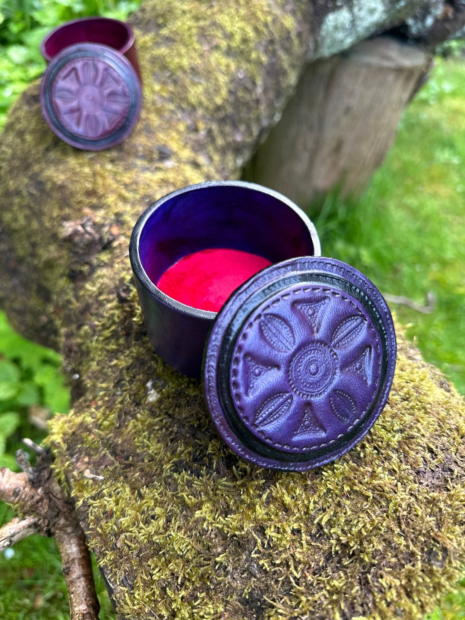 A Round African Tuareg Hand-Tooled Purple Leather Trinket/Jewellery Box by bibianna.co.uk with intricate designs and a red interior sits open on a mossy branch; another similar box rests amid lush green grass in the background.
