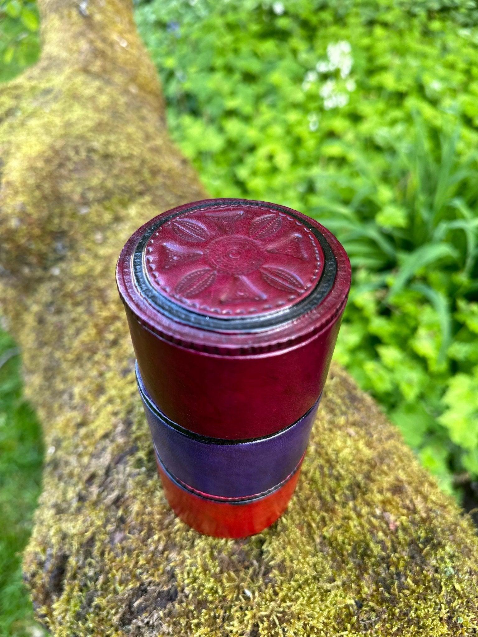 The Round African Tuareg Hand-Tooled Purple Leather Trinket/Jewellery Box by bibianna.co.uk, featuring a floral pattern on top, sits on a mossy tree trunk with green foliage blurred in the background.