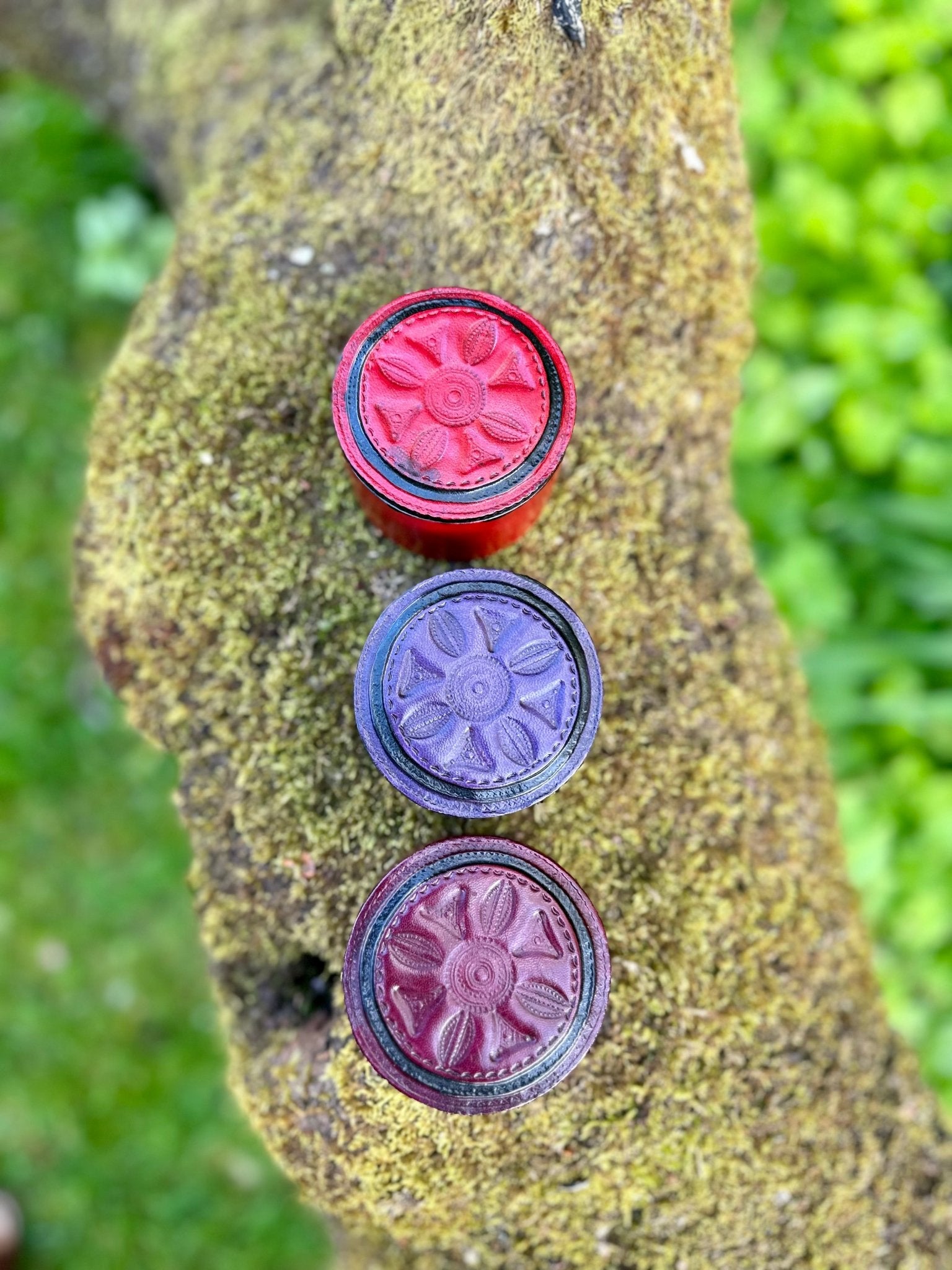 Three colored bottle caps—red, purple, and maroon—are arranged vertically on a mossy rock, resembling the vibrant lid of bibianna.co.uk’s Round African Tuareg Hand-Tooled Red Leather Trinket/Jewellery Box with grass in the background.