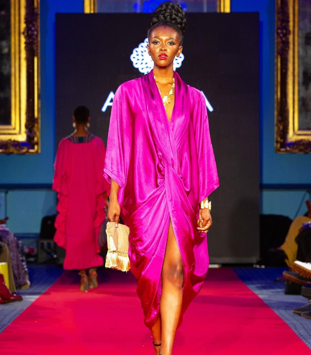 A model showcases the bibianna.co.uk Silk Dress—a bright pink, silky wrap with a thigh-high slit—while holding a fringe clutch. The backdrop displays gold-framed mirrors and a black screen.
