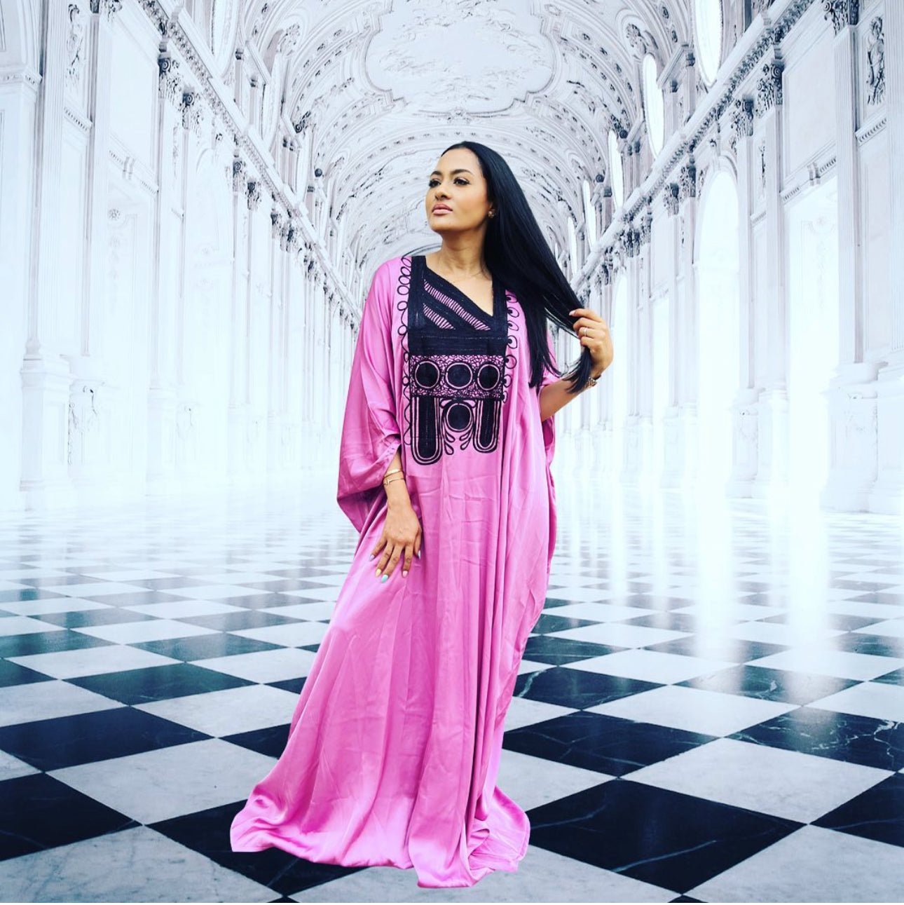 Wearing the Silk Embroidered Bubu from bibianna.co.uk, a woman stands confidently on a black-and-white checkered floor in an elegant hall with intricate white architecture and high ceilings.