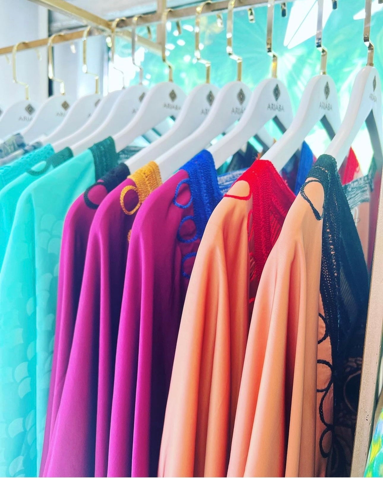 A close-up of vibrant Silk Embroidered Bubu dresses from bibianna.co.uk in teal, purple, blue, red, and peach on white hangers and a gold rack, creating a stylish ensemble.