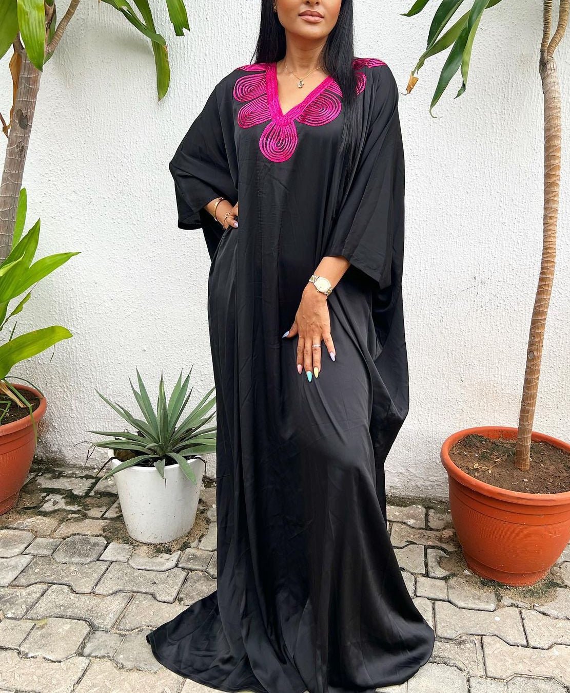 A woman stands outdoors in the Silk Embroidered Bubu - Fushia by bibianna.co.uk, featuring bright pink neckline detailing, surrounded by potted plants on a stone pavement before a white wall.