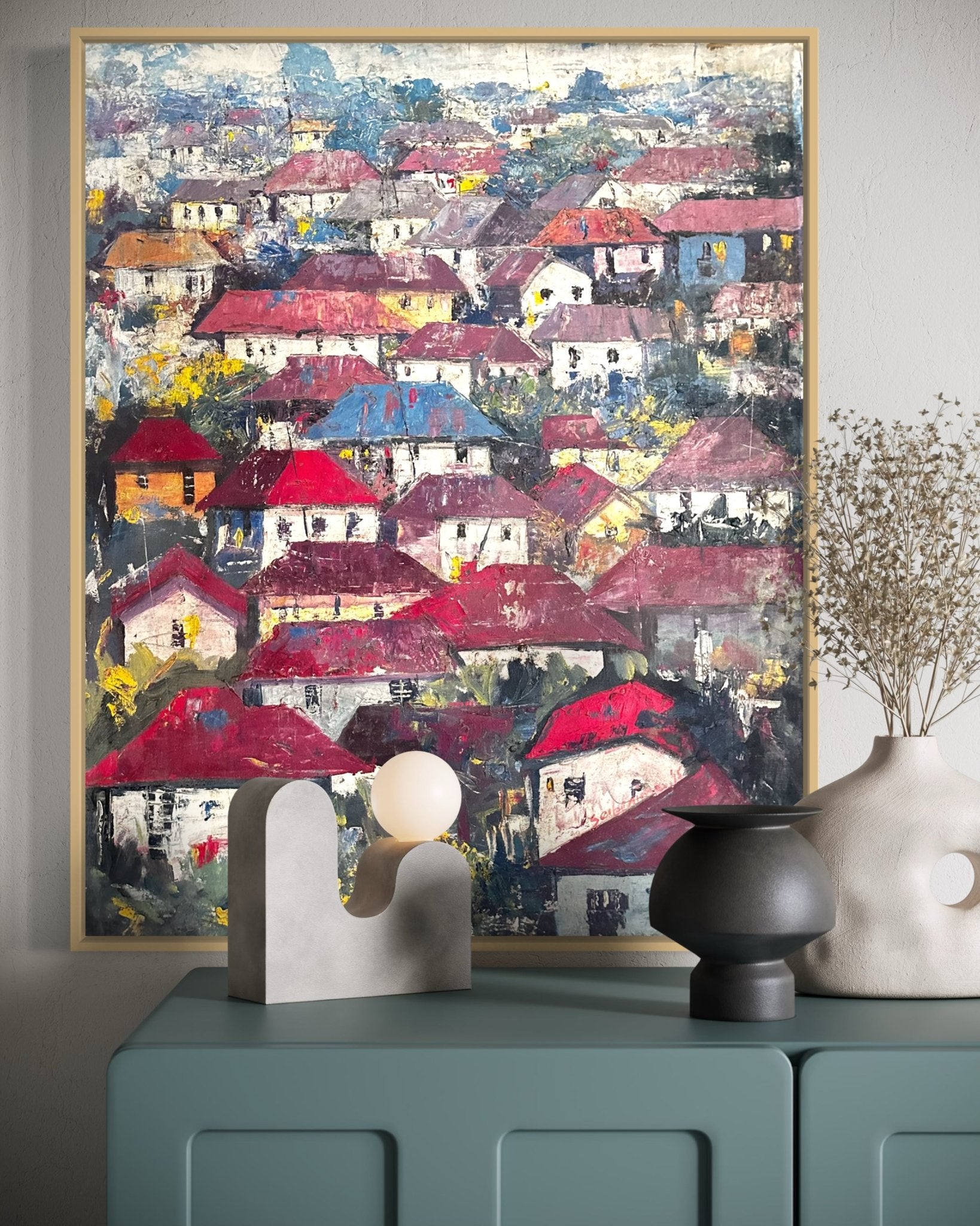Somewhere in the Africa. Rooftops" by Bibianna African Art Collective—a vibrant painting of a village with red and brown roofs—hangs above a blue cabinet accented with modern vases and dried flowers, enhancing its unique roofscape charm.