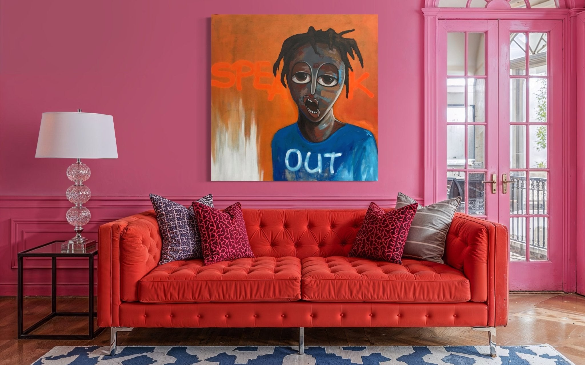 A vibrant living room with pink walls features a red tufted sofa, patterned pillows, and a glass lamp. Above the sofa hangs "Speak out," a hand-signed Achike Anayo painting from the United Kingdom artwork collection.