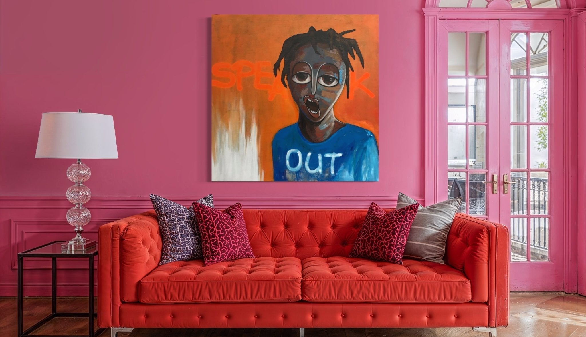 A vibrant living room with a tufted red sofa, patterned pillows, a modern glass lamp on a side table, and Achike Anayo’s hand-signed “Embracing The Smart & Digital Life” painting on a pink wall beside tall windows with pink trim.